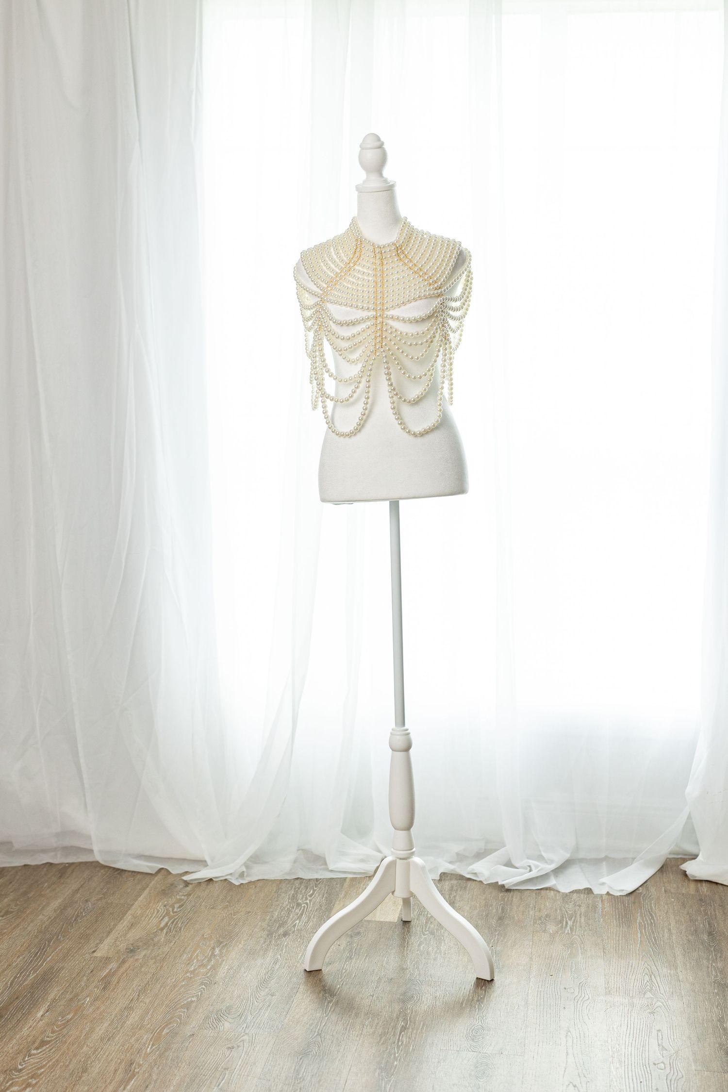 White mannequin torso on stand adorned with elegant pearl body chain necklace, displayed against sheer white curtains.