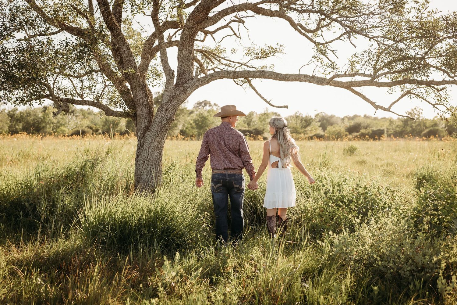Couple walking hand in hand through tall grass in a golden sunlit field by a tree.