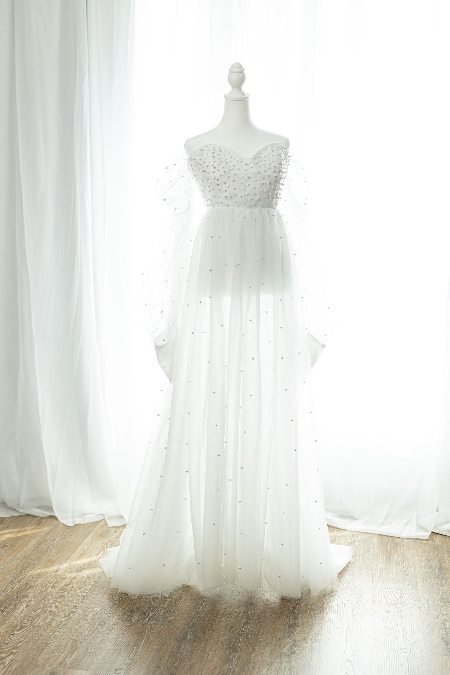 Elegant white tulle wedding gown with pearl embellishments displayed on a mannequin near sheer curtains.