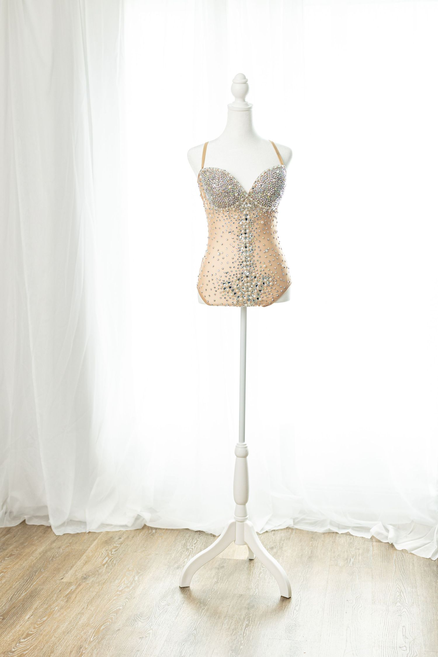 Elegant rhinestone-embellished nude bodysuit displayed on a white mannequin stand against sheer white curtains.