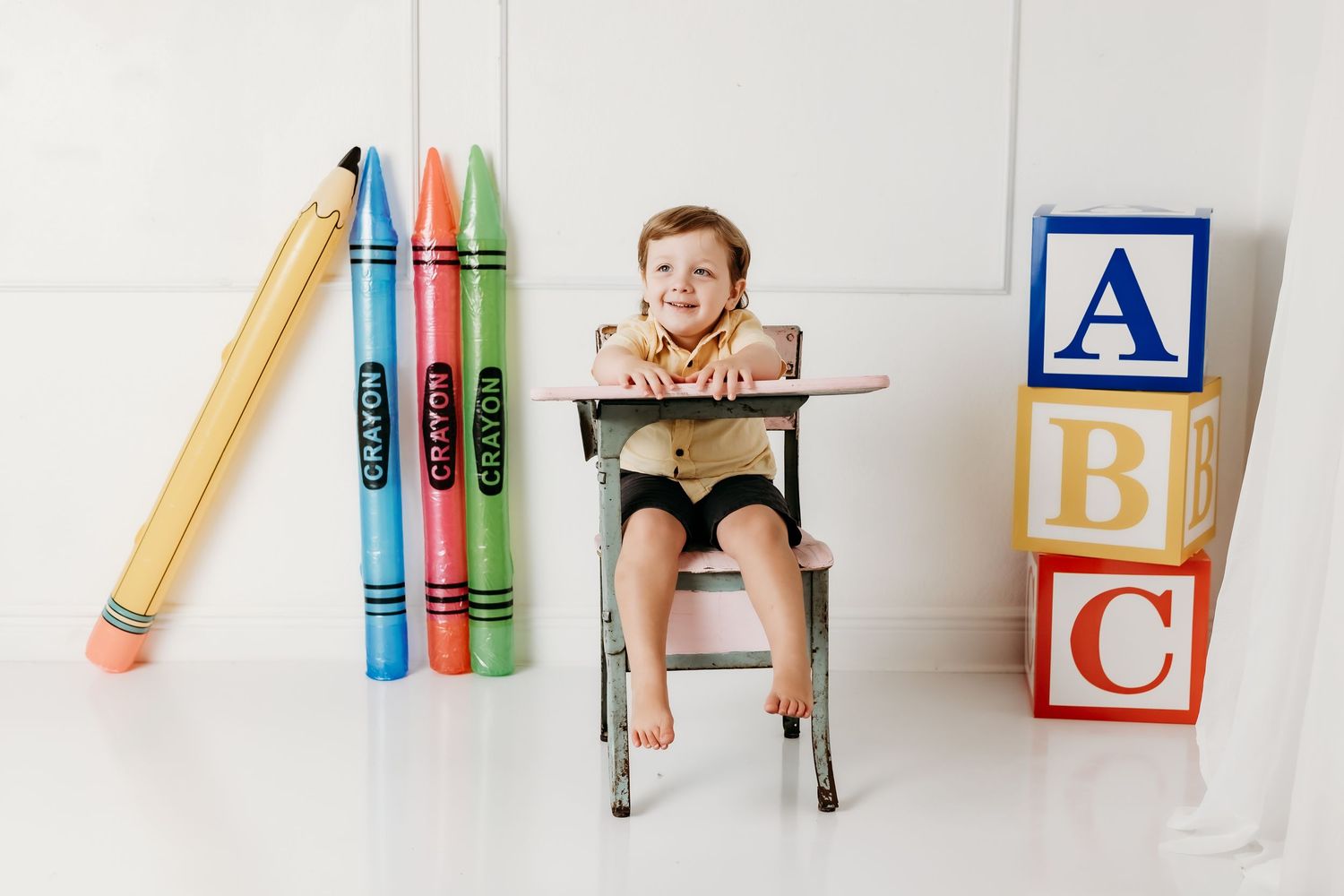 Cheerful boy sits at antique pink school desk flanked by oversized crayon props and alphabet blocks.