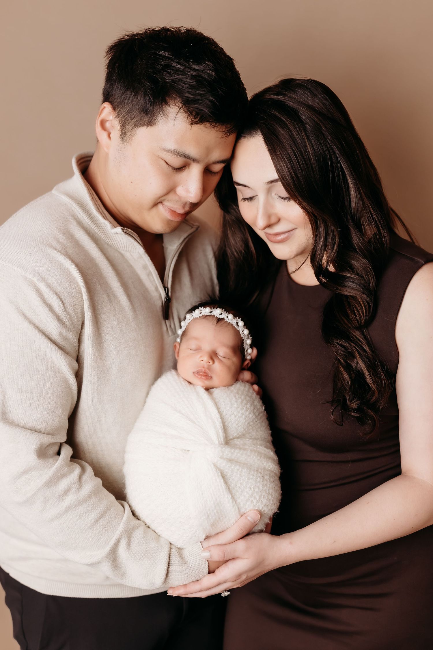 Parents tenderly hold their swaddled newborn baby with floral headband in a warm studio portrait.