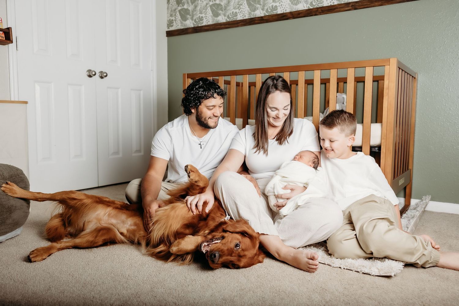 Family with toddler and newborn baby sitting on nursery floor with golden retriever dog.