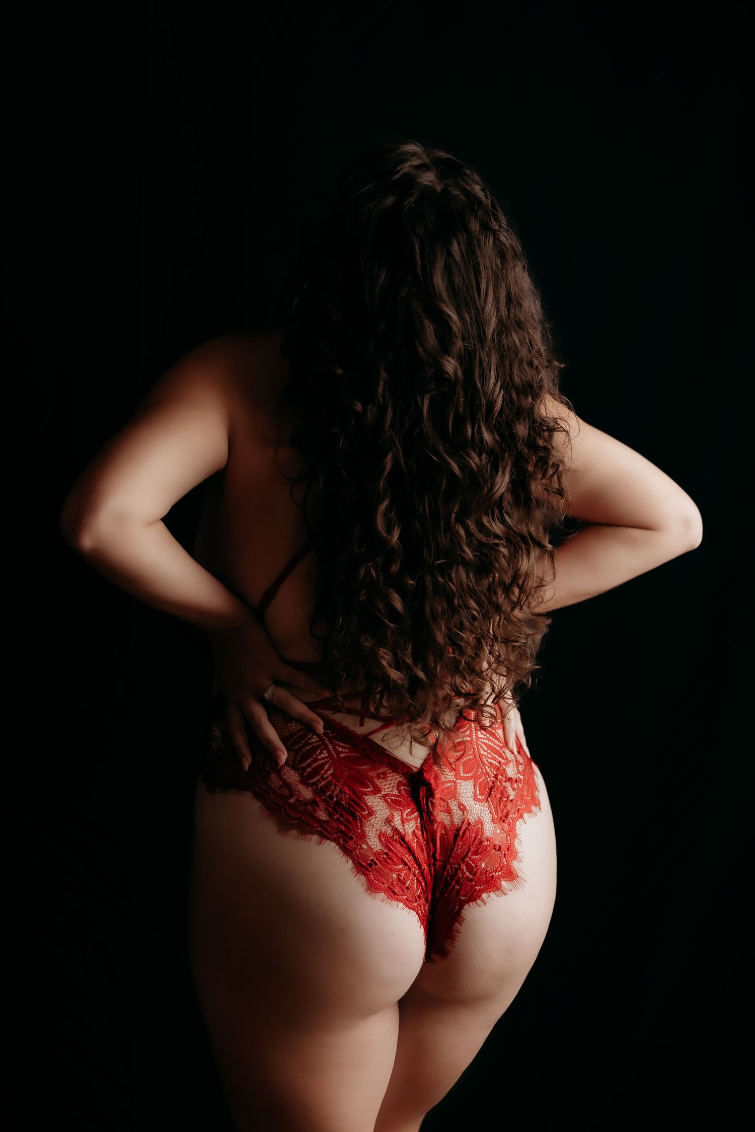 Woman with curly hair in red lace bodysuit posing from behind on dark background.