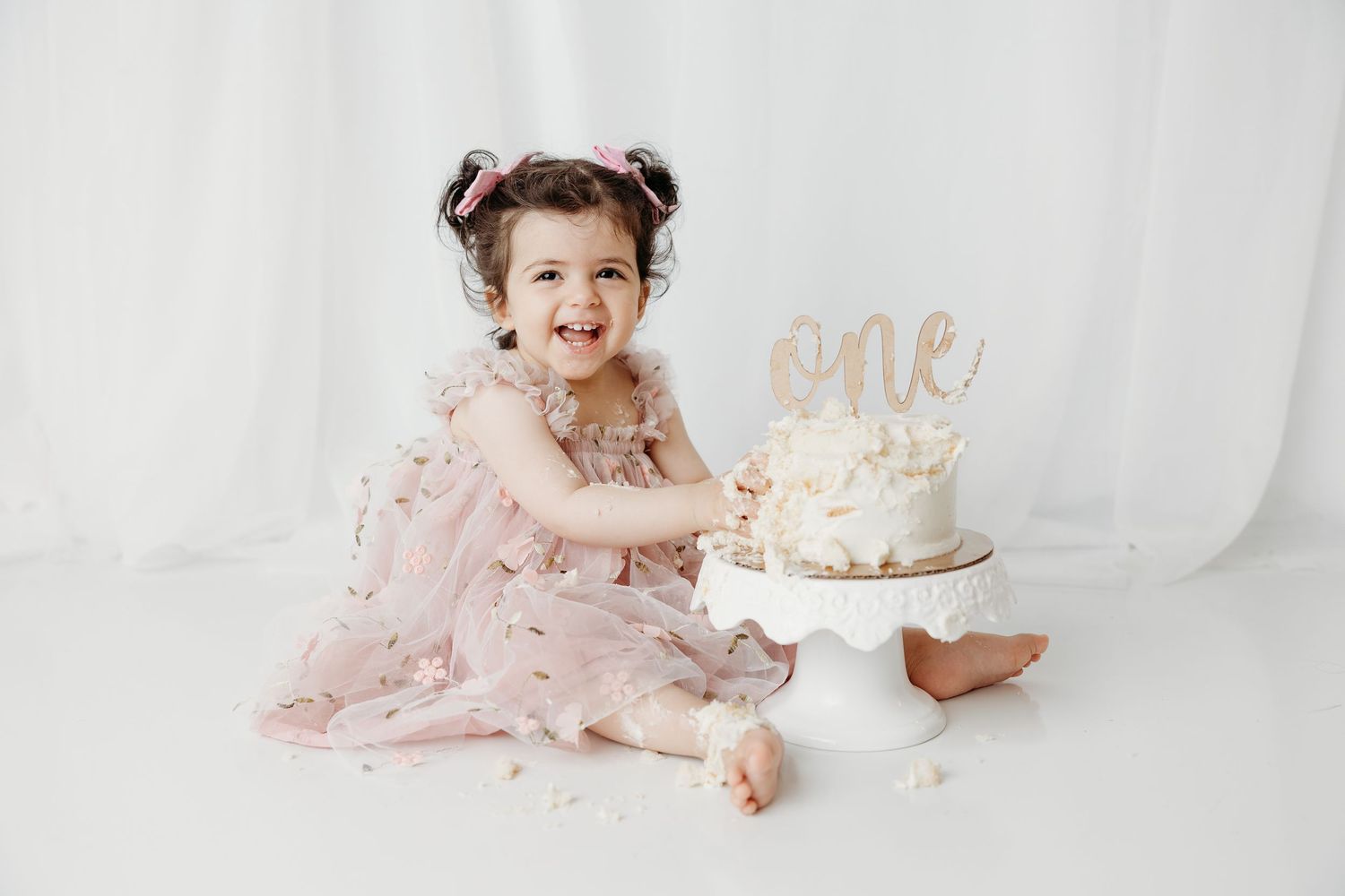 Smiling baby girl in pink tulle dress celebrates first birthday smashing a white cake with 'one' topper.