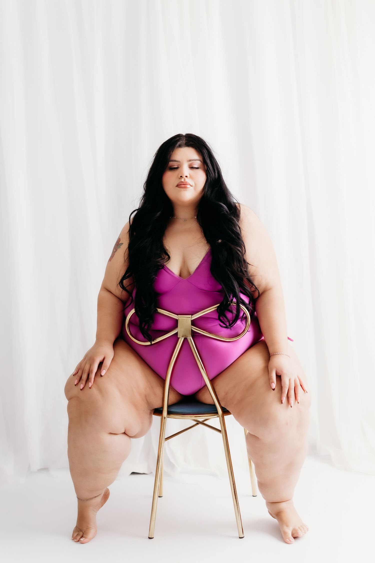 Plus size woman in purple bodysuit sitting confidently on gold chair against white curtain.