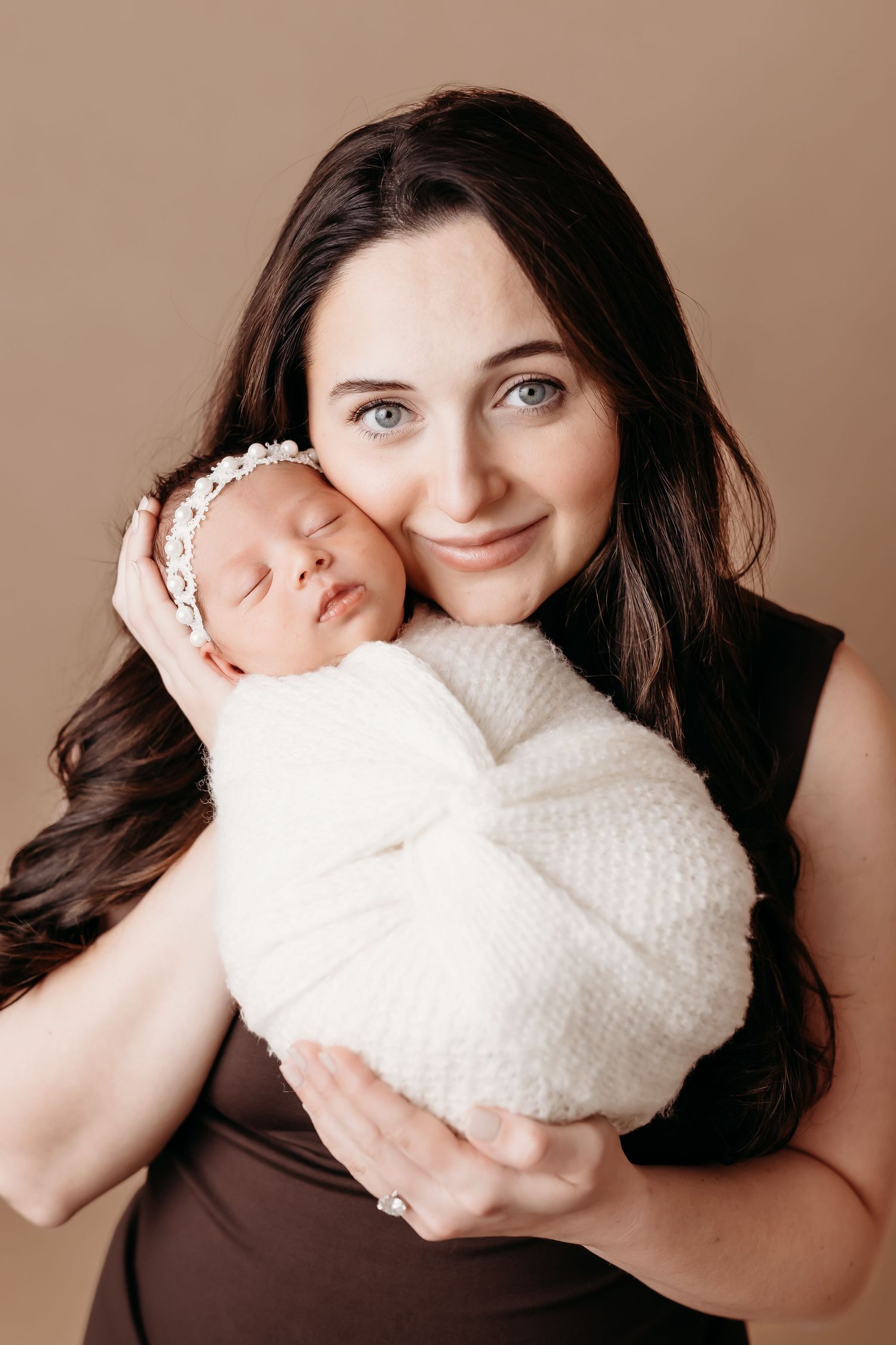 A smiling dark-haired mother holds her sleeping newborn baby swaddled in white with a floral headband.