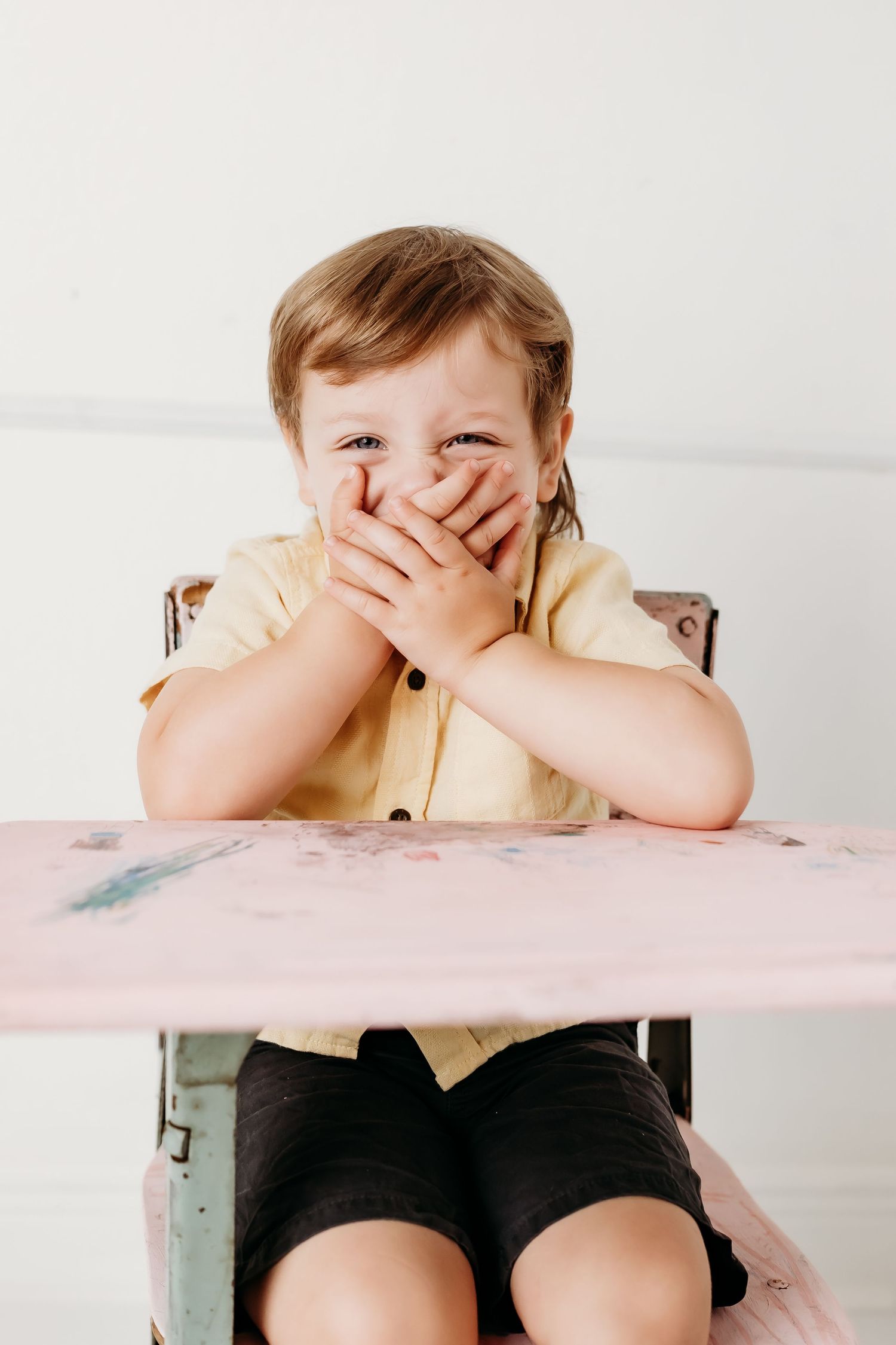 Laughing toddler boy covers mouth with hands while sitting at a vintage pink school desk.