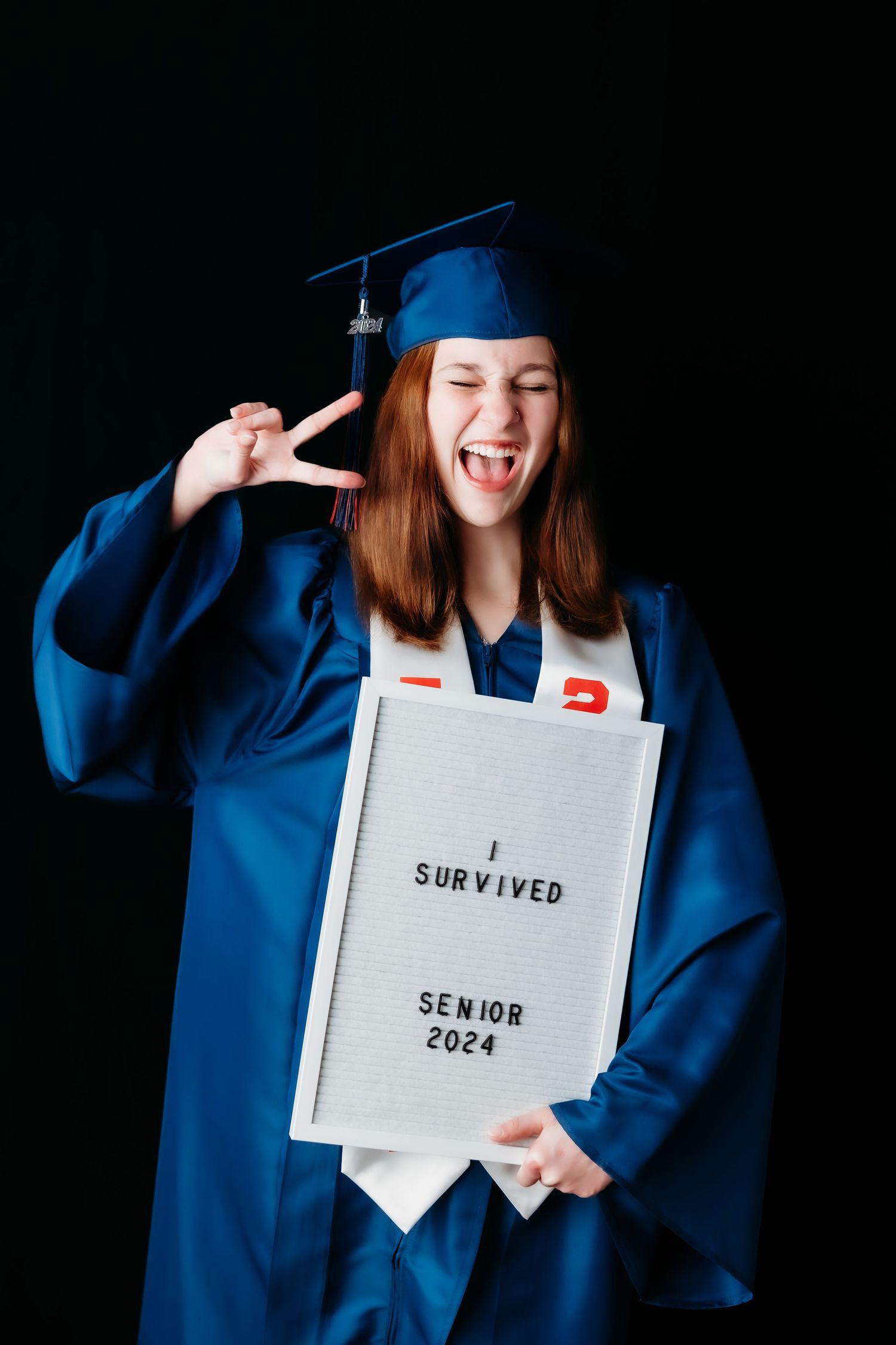 Excited graduate in blue cap and gown holding sign reading I Survived Senior 2024.