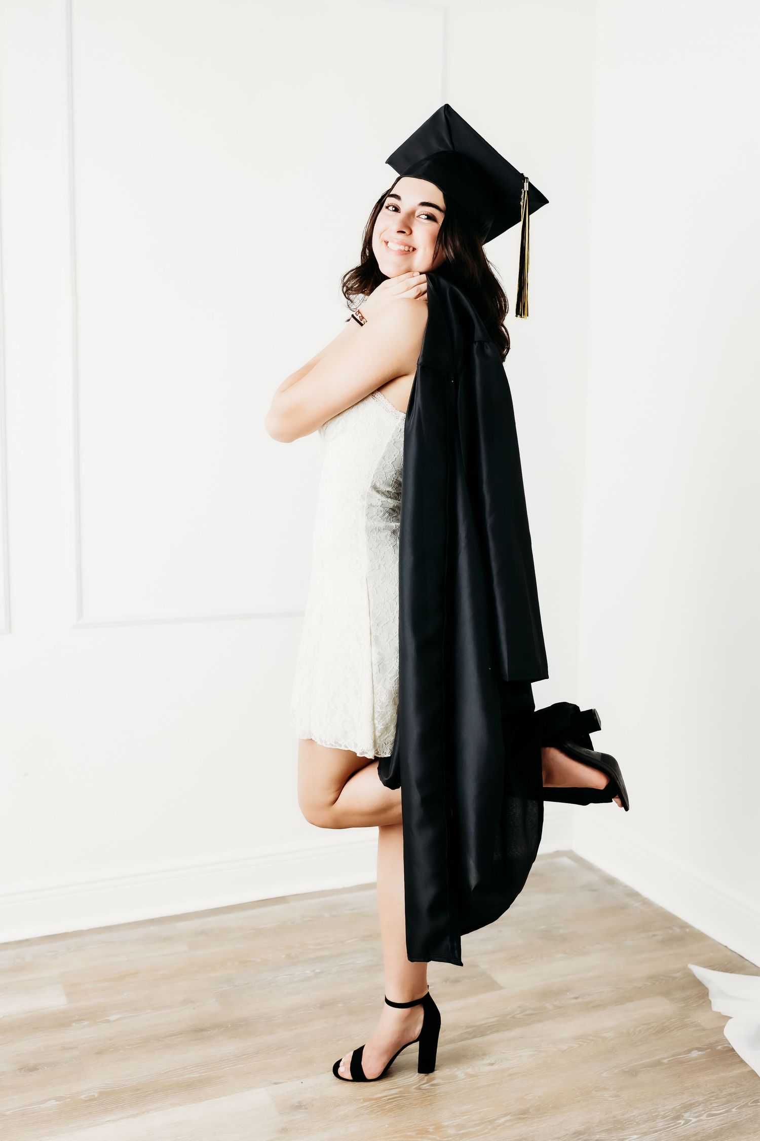 Brunette graduate in black cap holding gown over white dress indoors on hardwood floor.