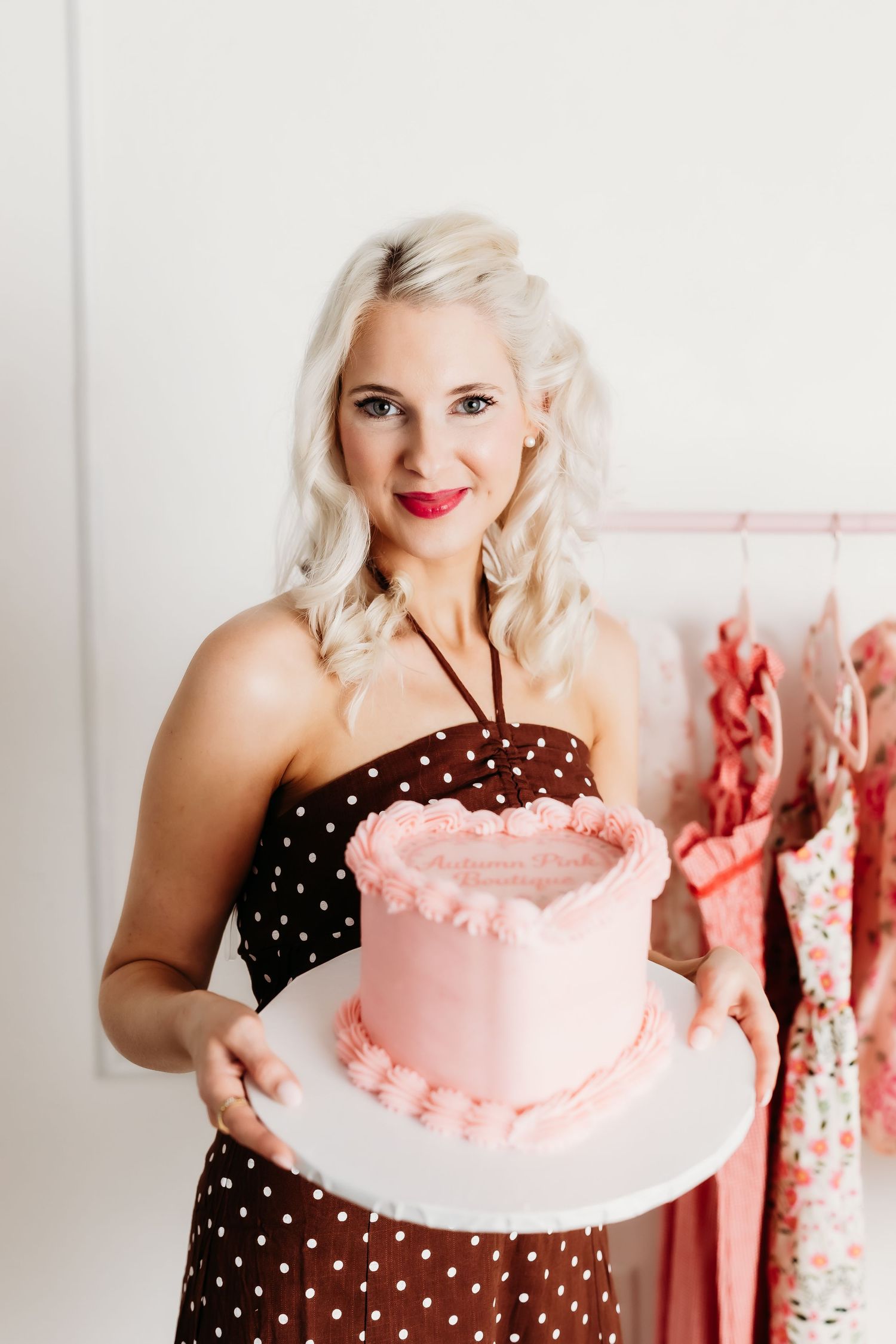 Smiling blonde woman in polka dot dress holding a pink frosted celebration cake.
