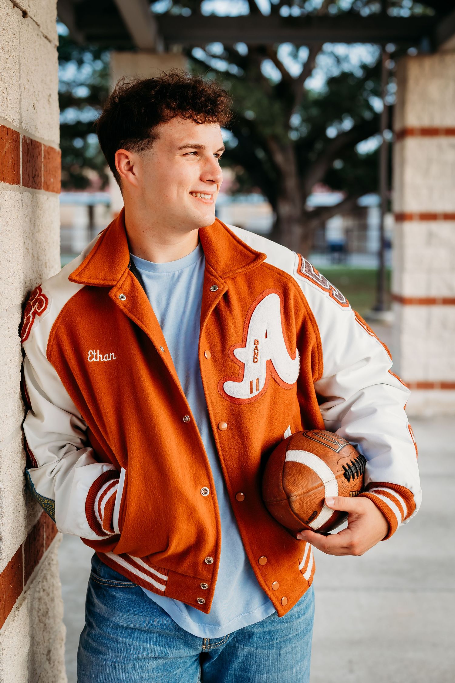 Senior boy named Ethan in orange letterman jacket holds football leaning against brick wall.