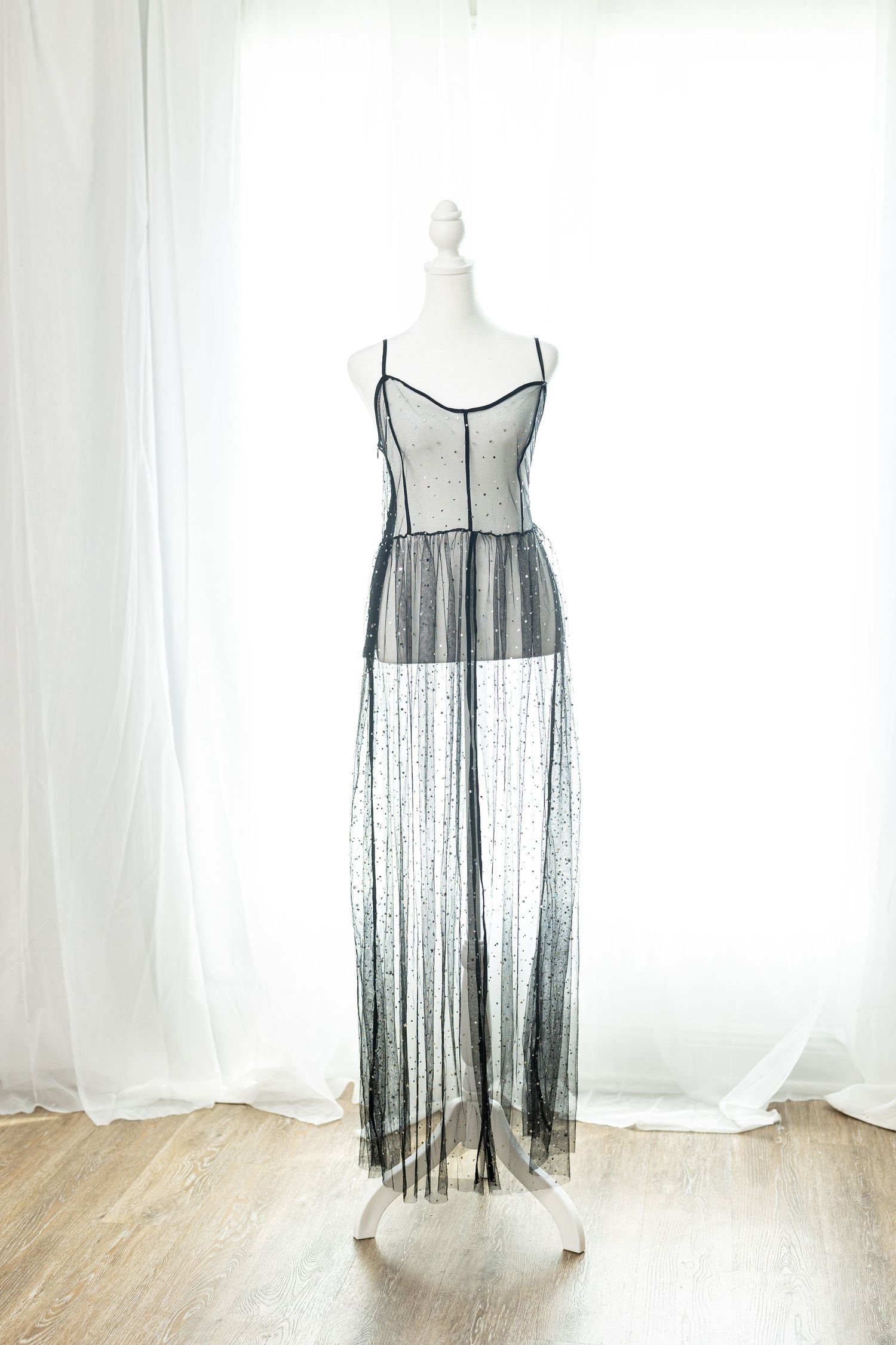 Elegant sheer black lace maxi dress with spaghetti straps displayed on a white mannequin near curtains.