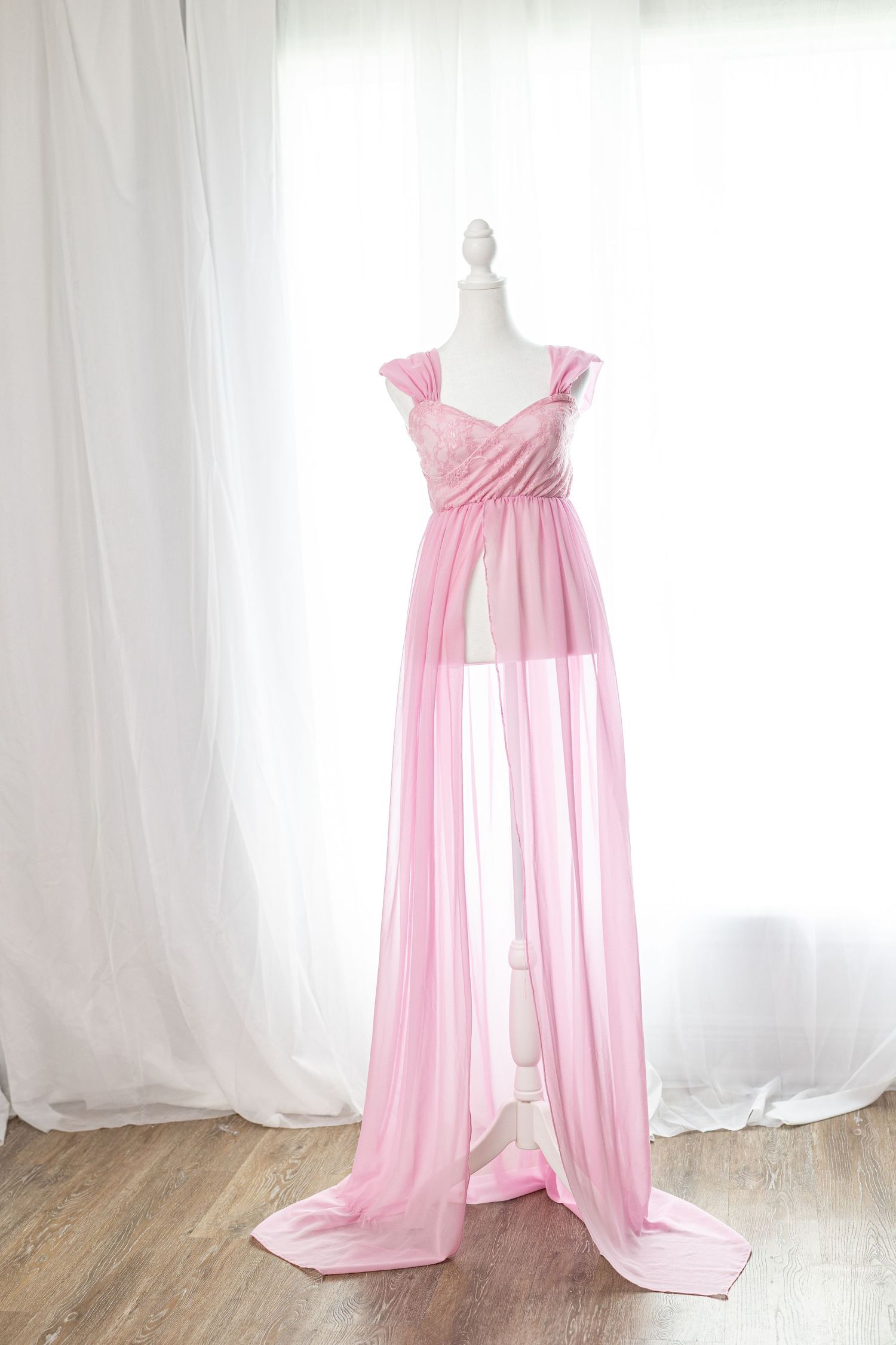 Pink chiffon maternity gown on mannequin with lace bodice and flowing sheer skirt, displayed against white curtains.
