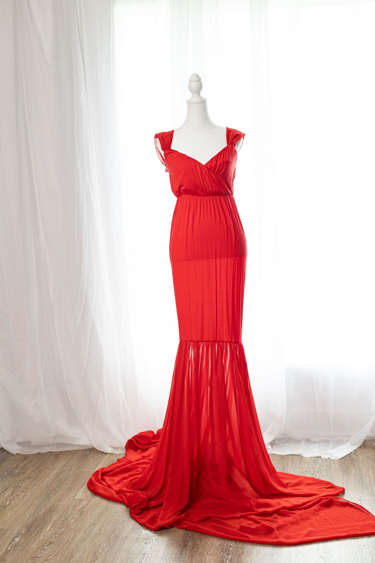 Elegant red mermaid gown with ruffle straps and long train displayed on a mannequin against white curtains.