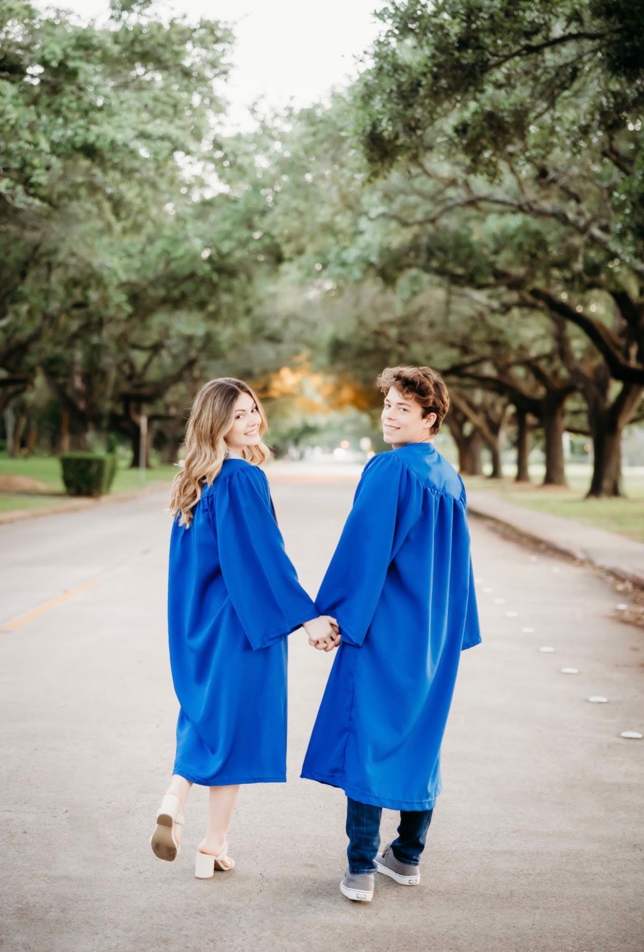 Two graduates in blue gowns hold hands walking down tree-lined path for senior photos.
