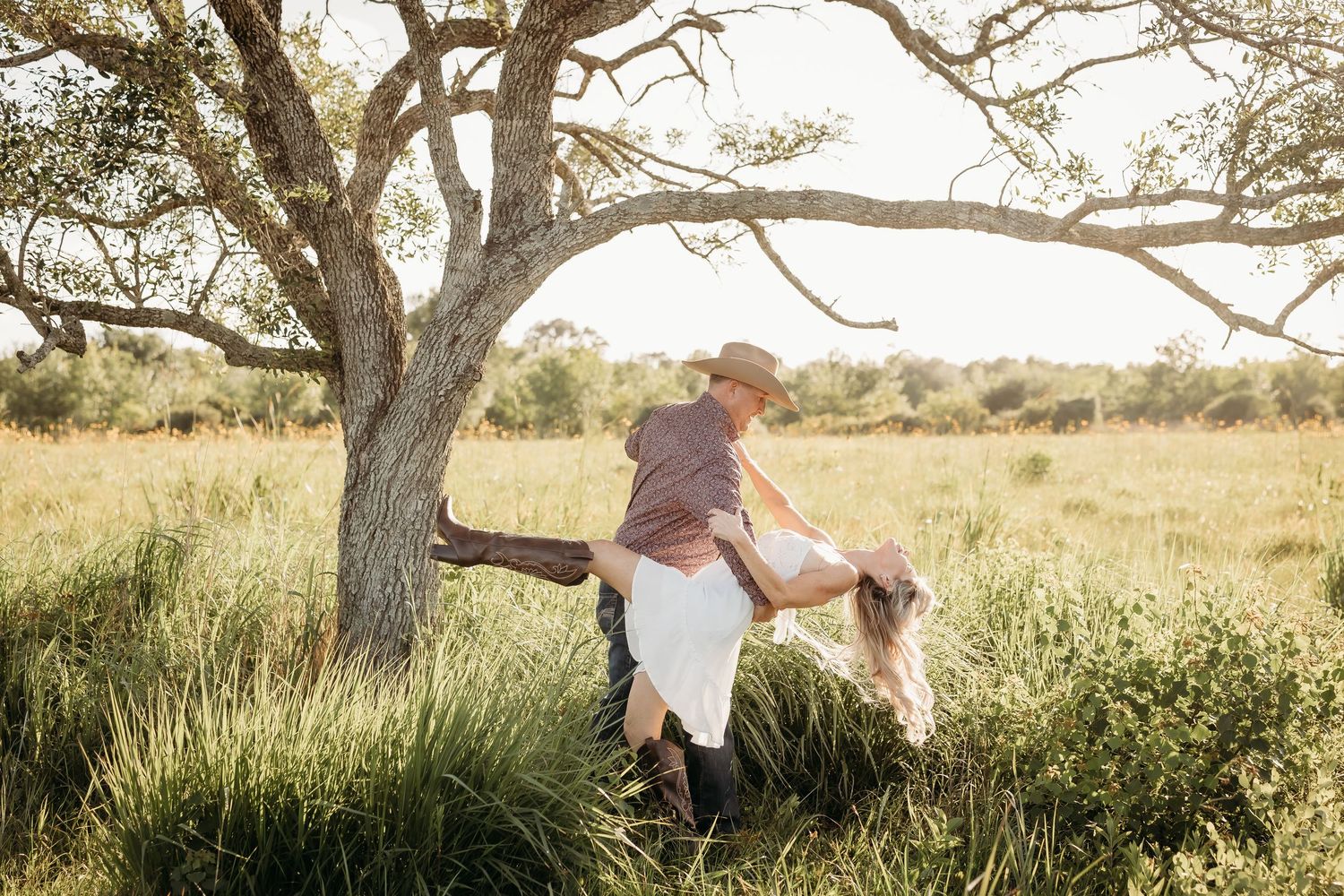 Couple dancing playfully in a sunlit grassy field beneath a sprawling oak tree.
