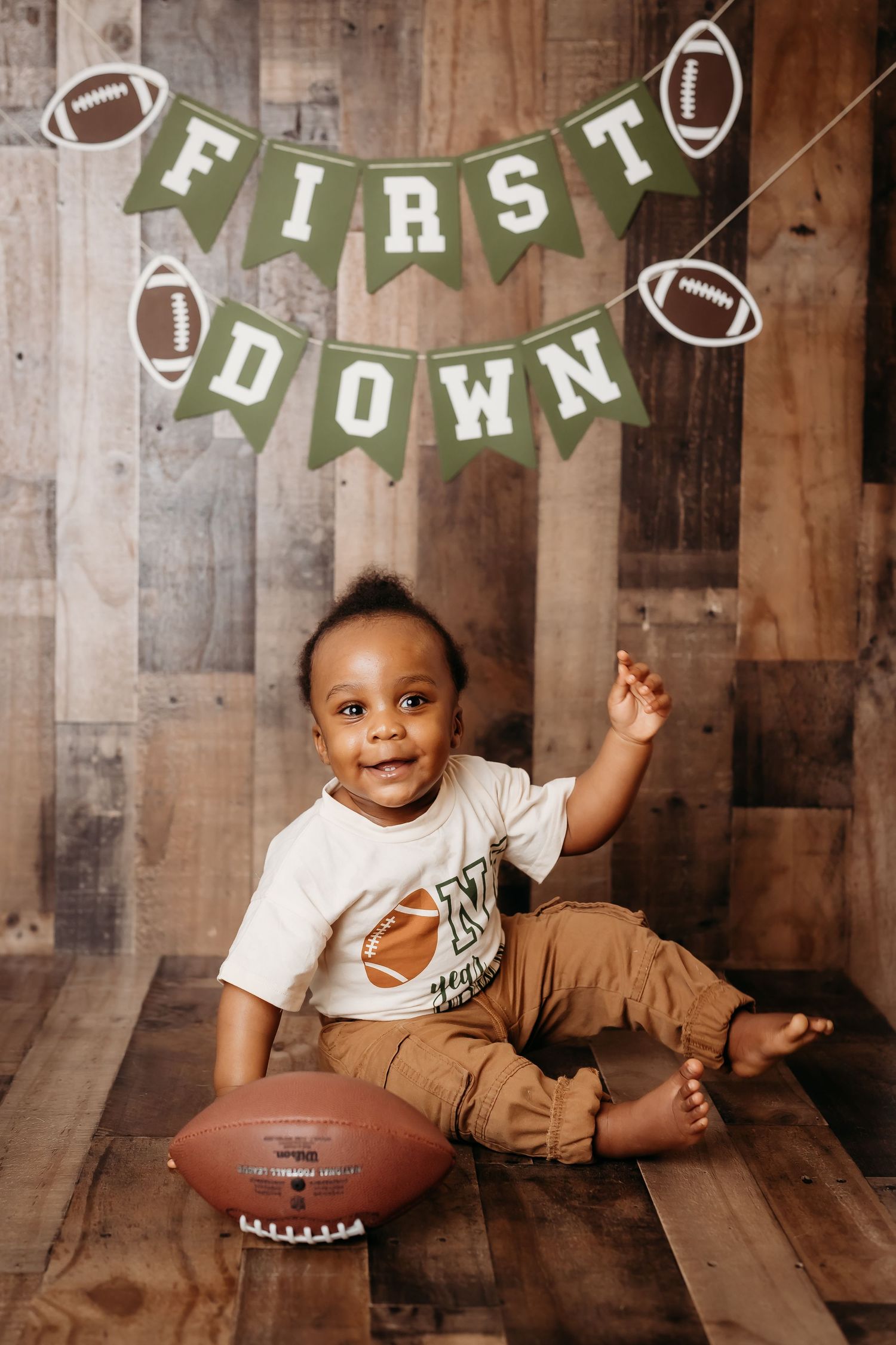 Baby boy sitting with a football under a First Down banner for his first birthday photo.
