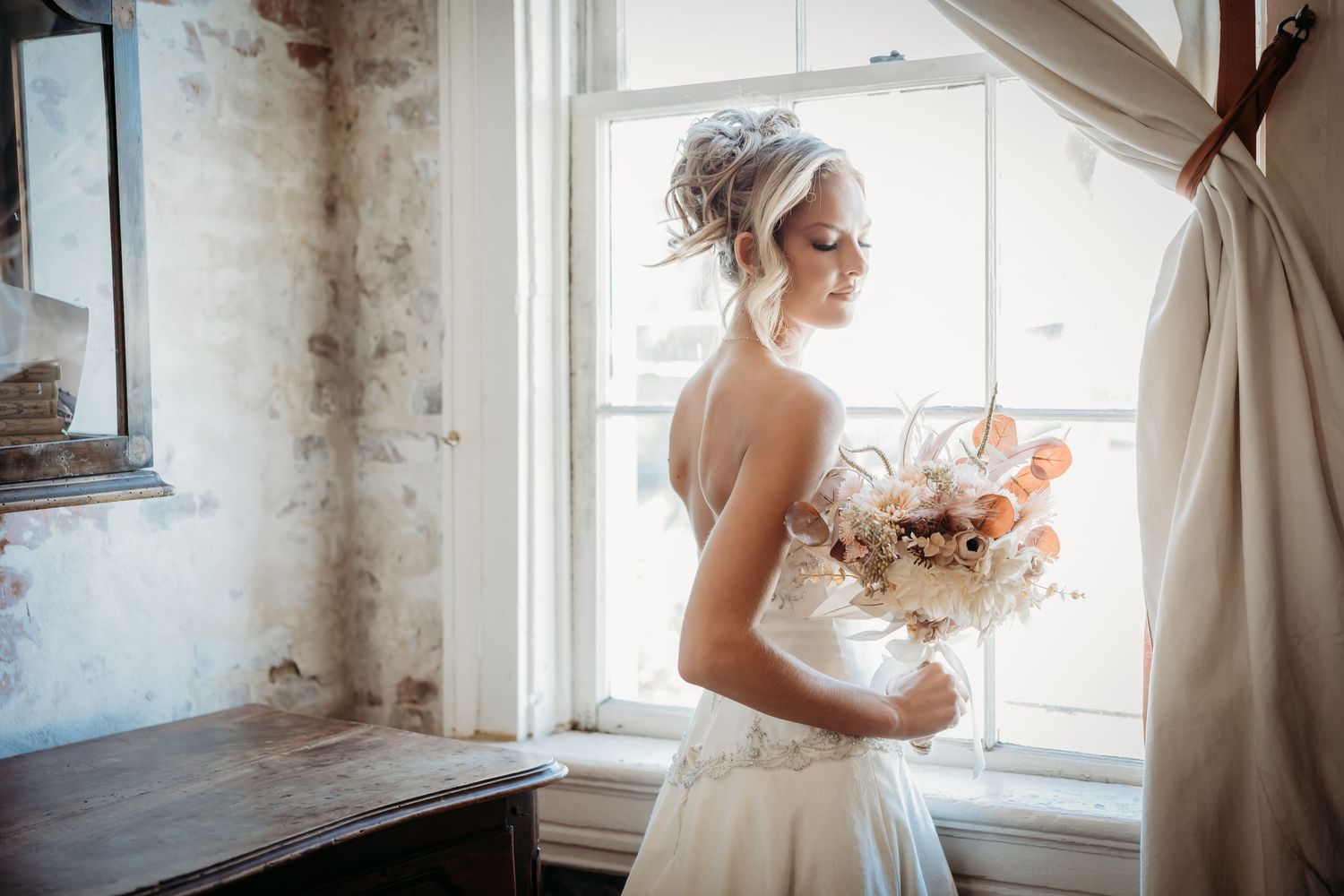 Elegant bride in strapless gown holds boho bouquet by vintage window in rustic venue.