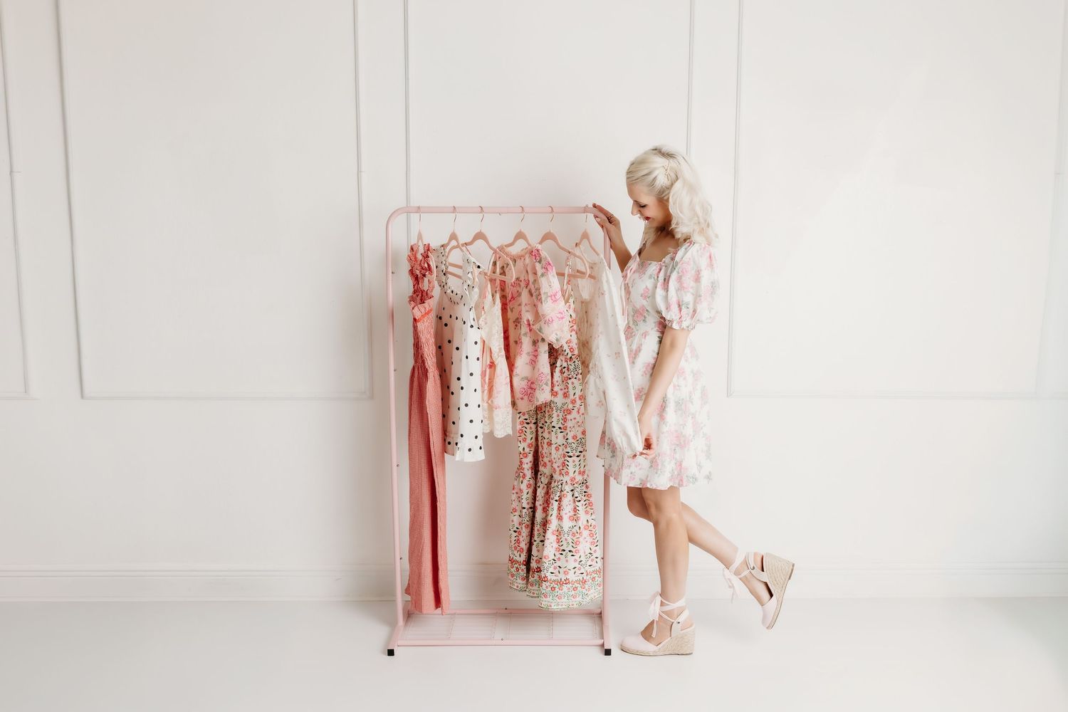 Blonde woman in white dress browsing pink floral dresses on a pink clothing rack.
