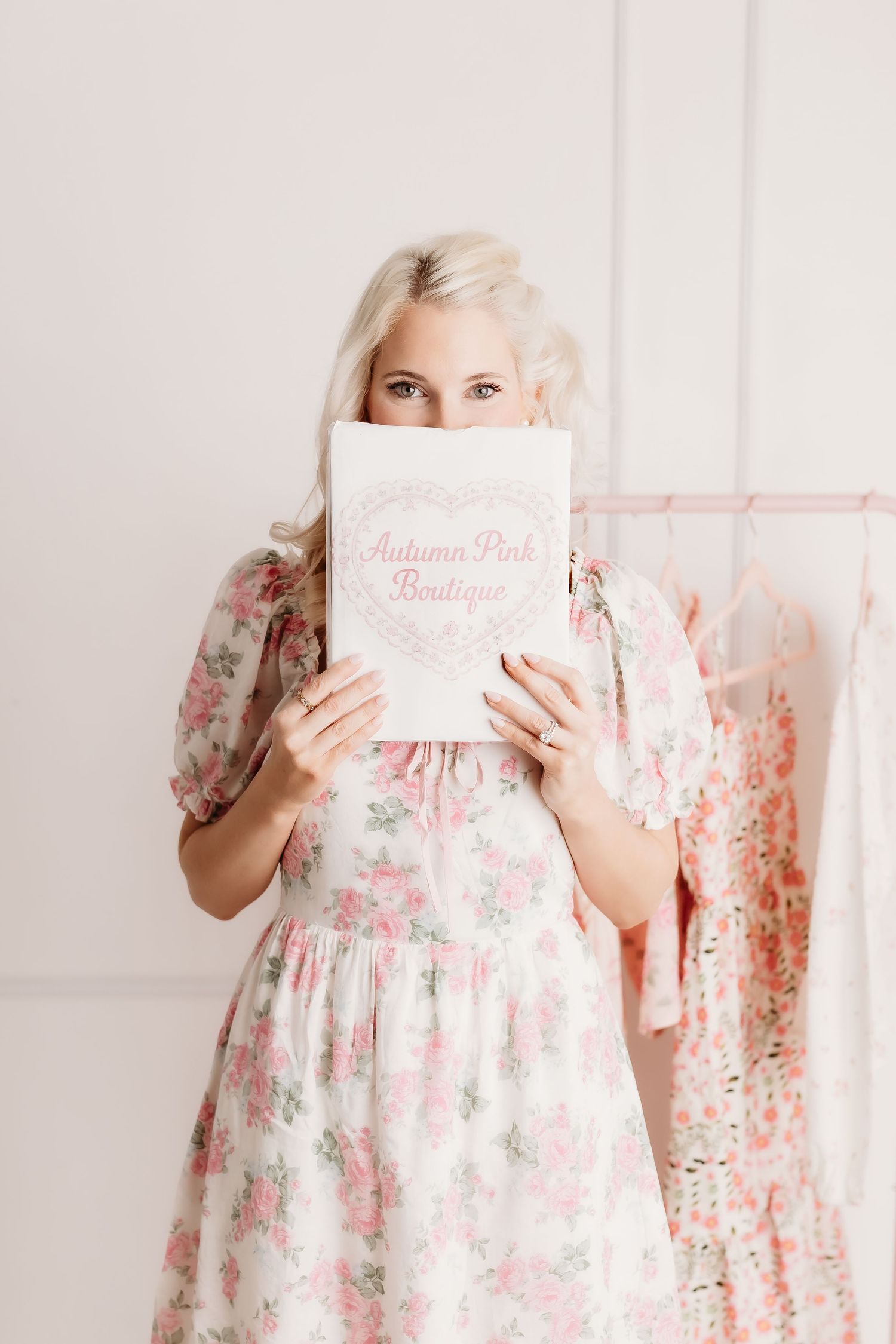 Blonde woman in floral dress playfully hiding behind Autumn Pink Boutique sign near clothing rack.