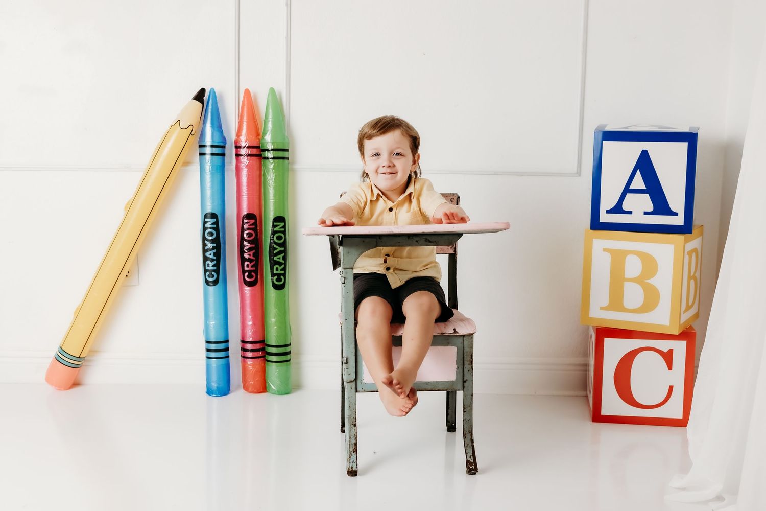 Young boy smiles at vintage pink school desk with giant crayon props and ABC blocks in white studio.