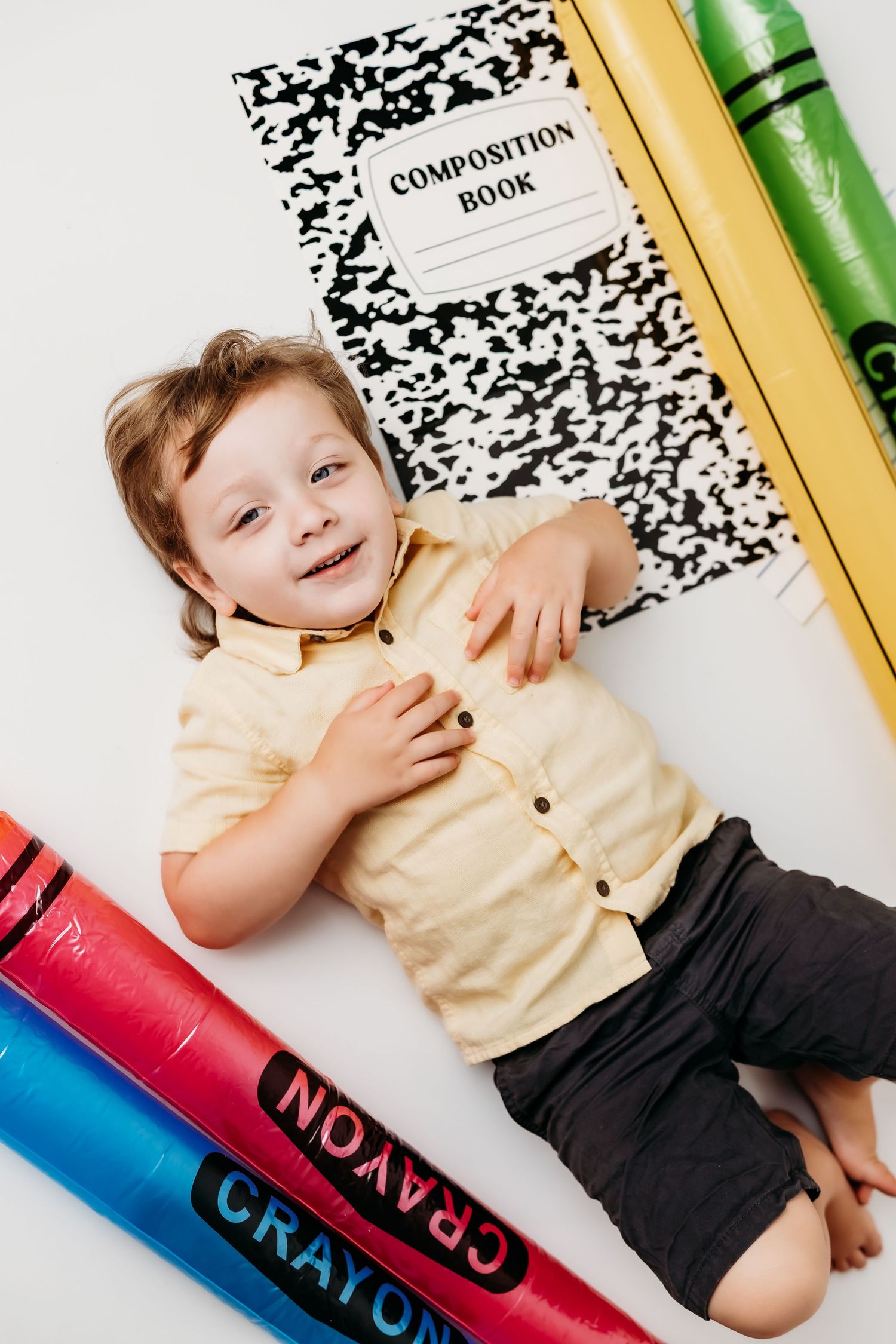 A happy young child in a yellow shirt lies on a white floor surrounded by oversized crayons and a composition book.