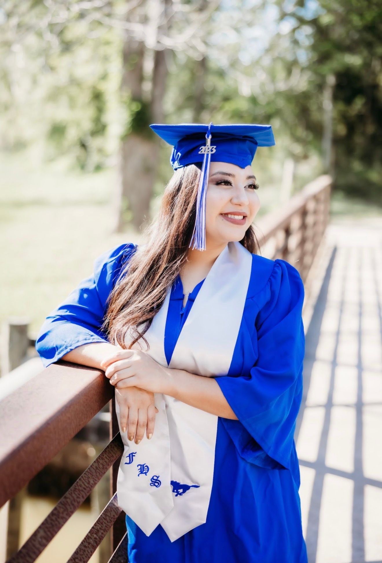 Smiling female graduate in blue cap and gown leans on wooden bridge railing outdoors.