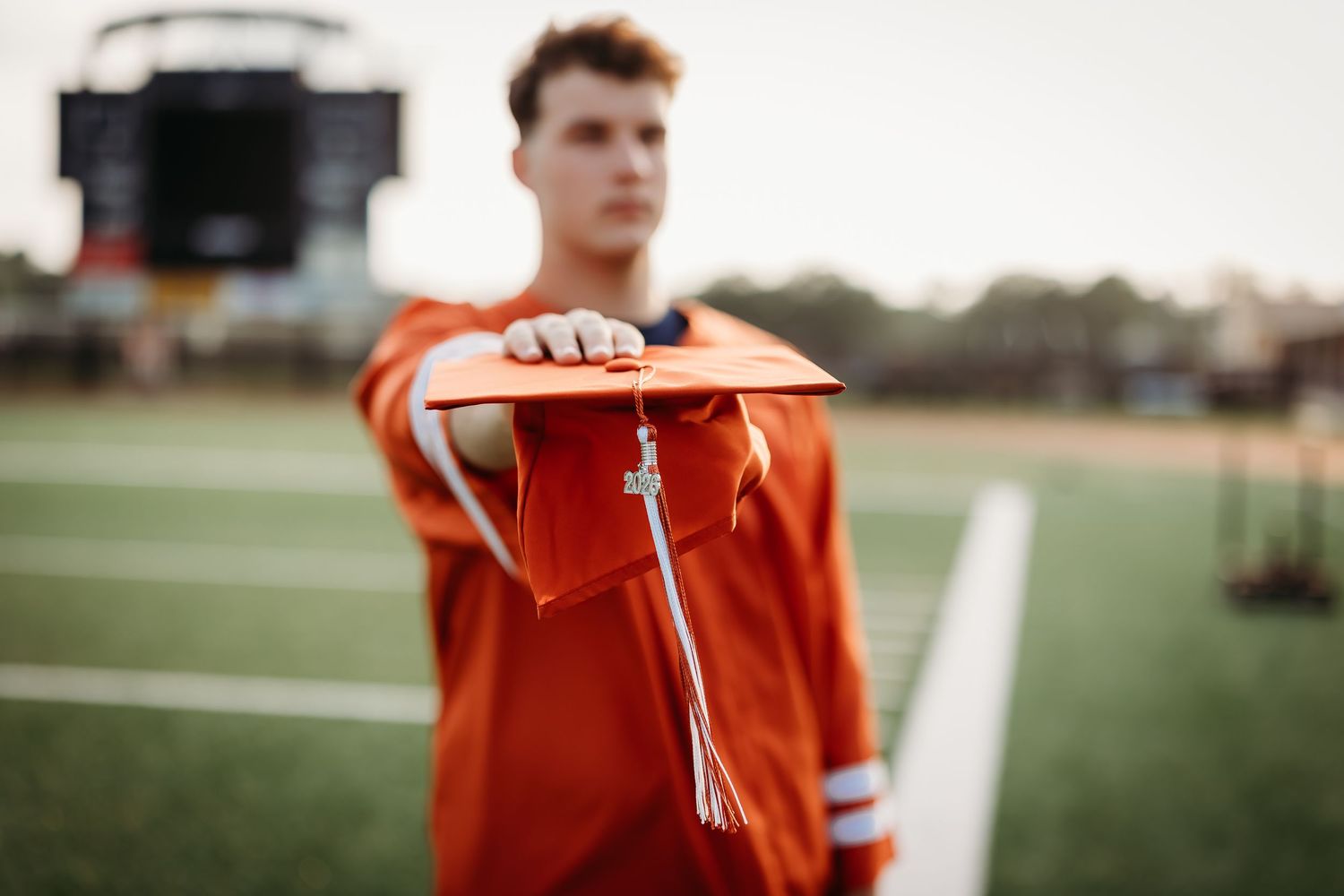 Male graduate in orange gown extending graduation cap toward camera on green football field.
