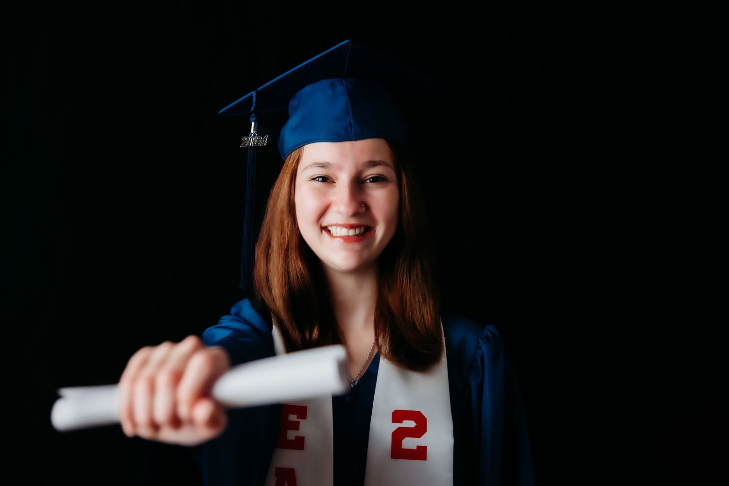 Smiling redhead graduate in blue cap and gown holding diploma against dark background.