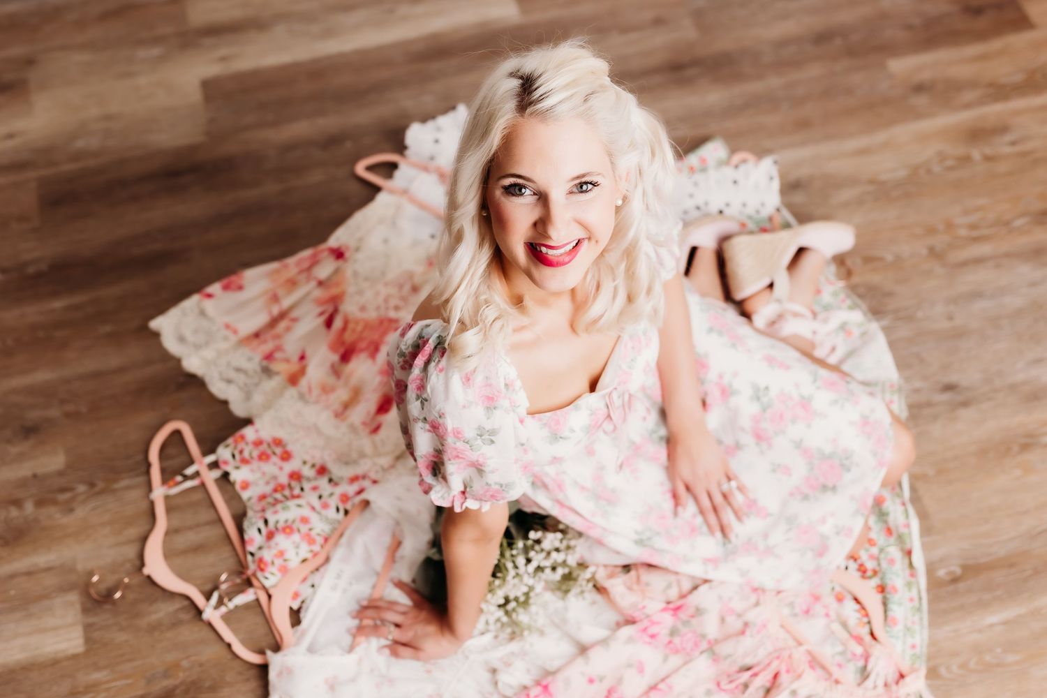 Smiling blonde woman in pink floral dress sitting on wooden floor surrounded by clothing and accessories.