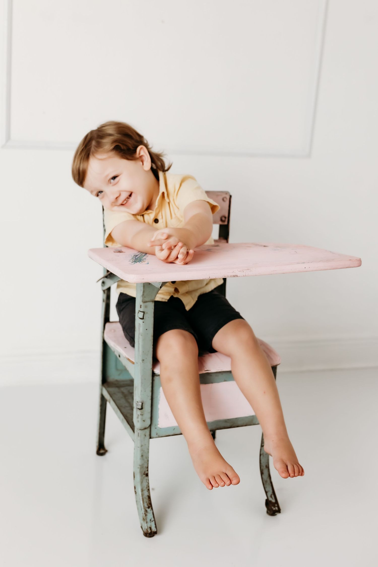 Barefoot toddler boy leans on vintage pink school desk smiling in bright white photography studio.
