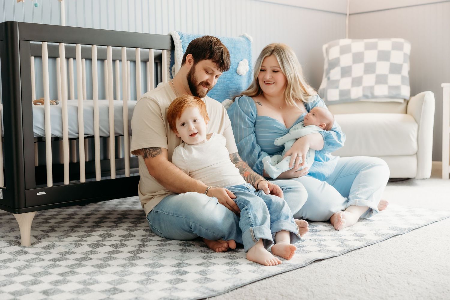 Happy family with toddler and newborn sitting together on nursery floor near crib.