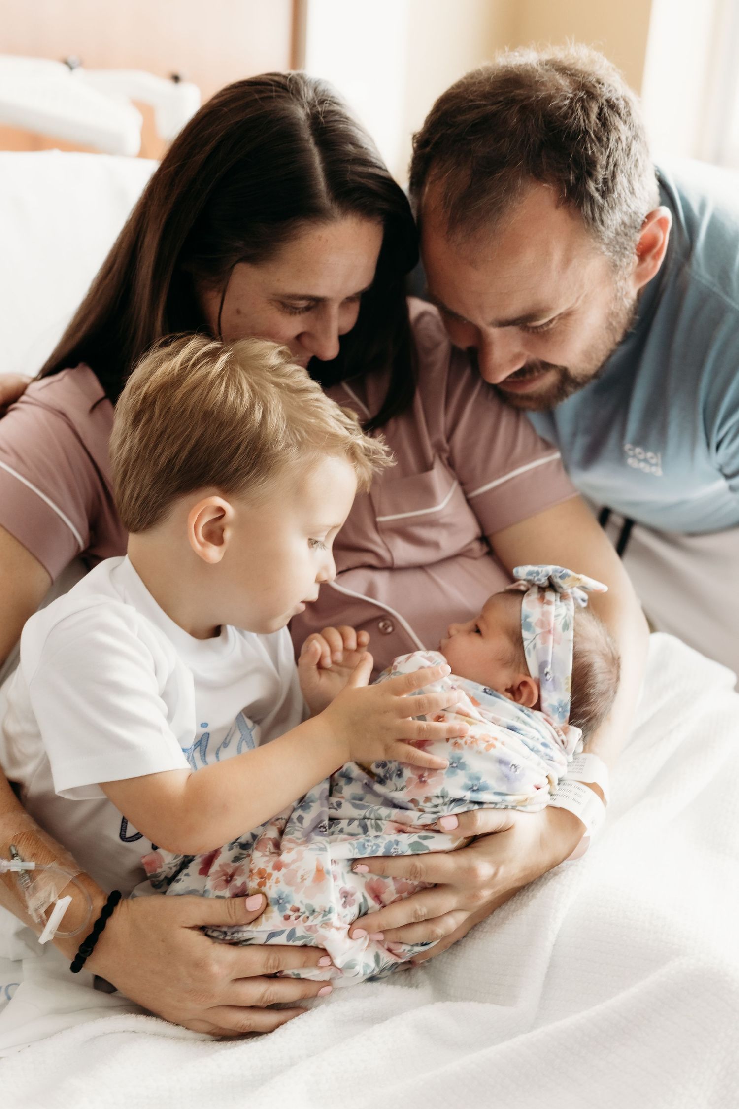 Family of four admiring their newborn baby, with parents and young boy gathered closely together on a hospital bed.