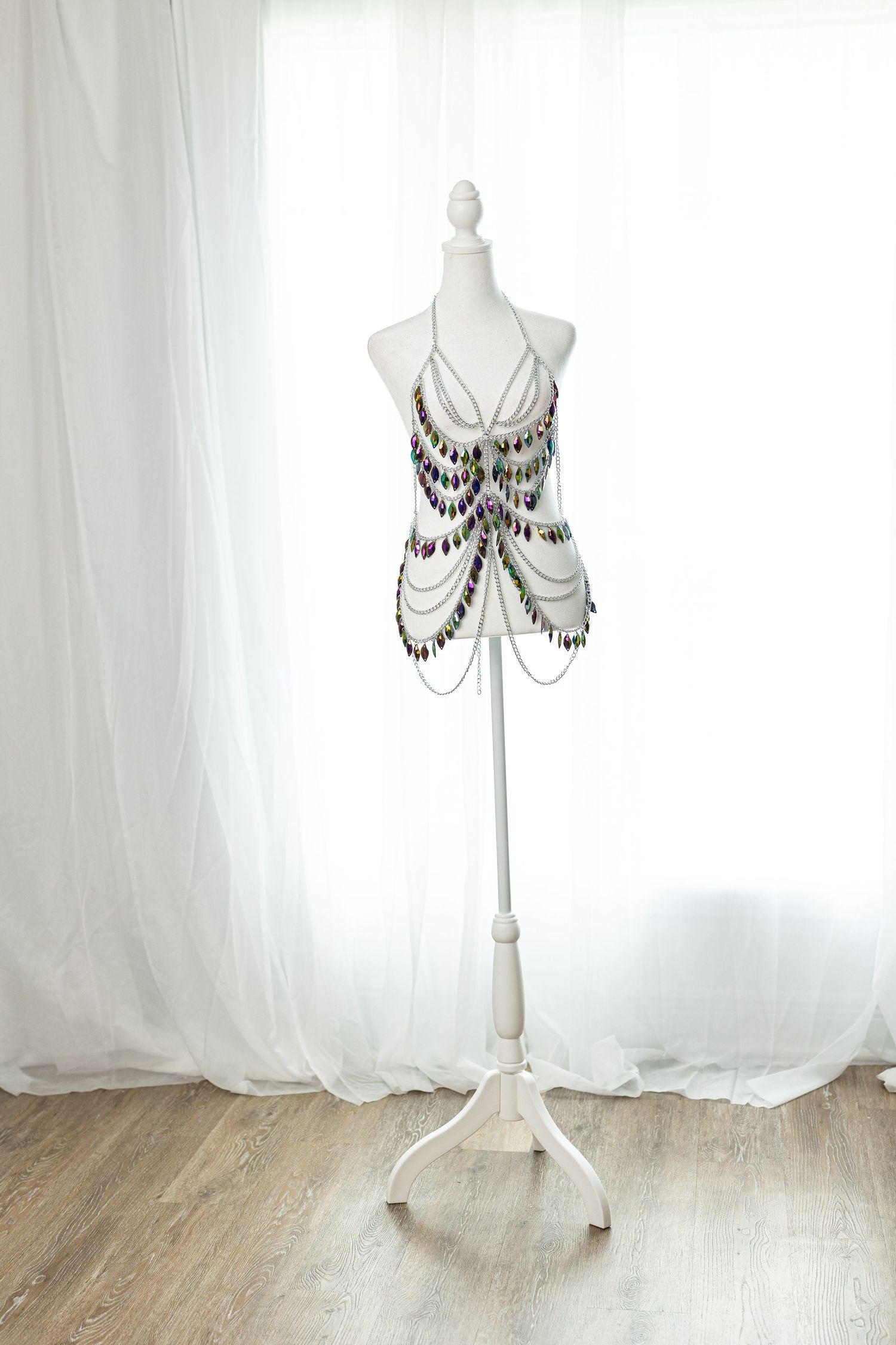 White mannequin display stand adorned with colorful gemstone and chain body jewelry against sheer white curtains.
