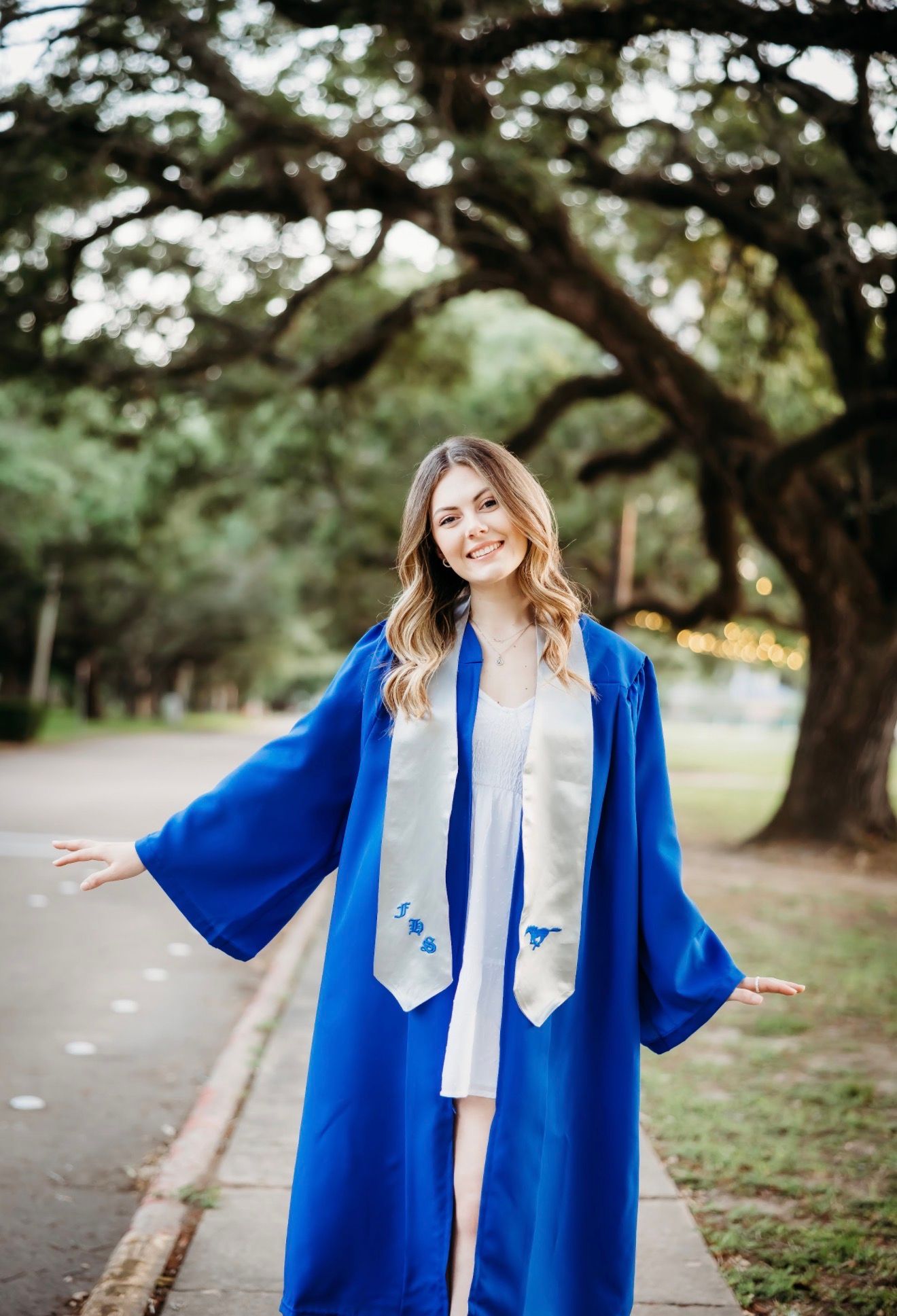 Smiling female graduate in royal blue gown with stole poses on scenic tree-lined path.