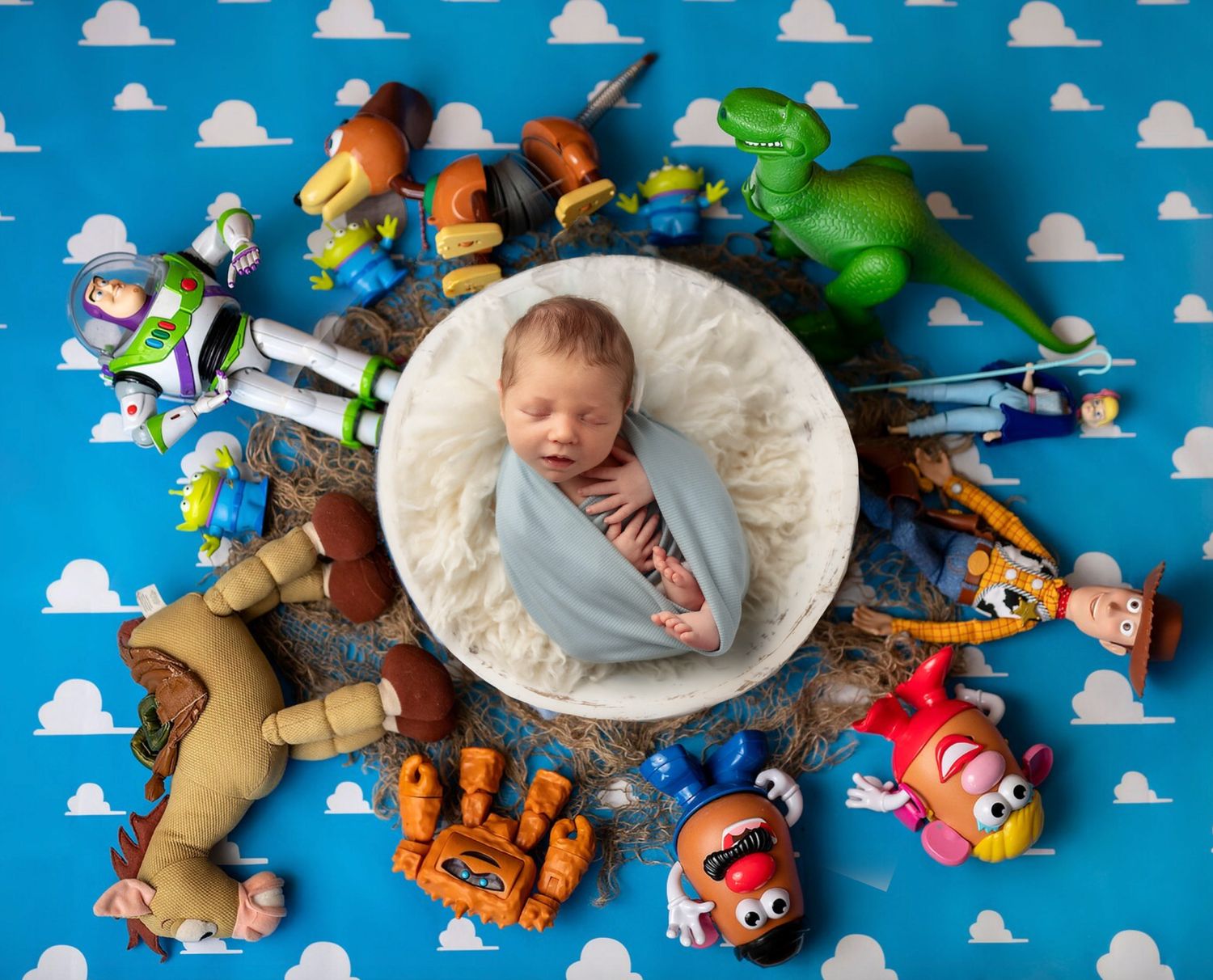 Sleeping newborn baby swaddled in blue wrap surrounded by colorful Toy Story character toys.