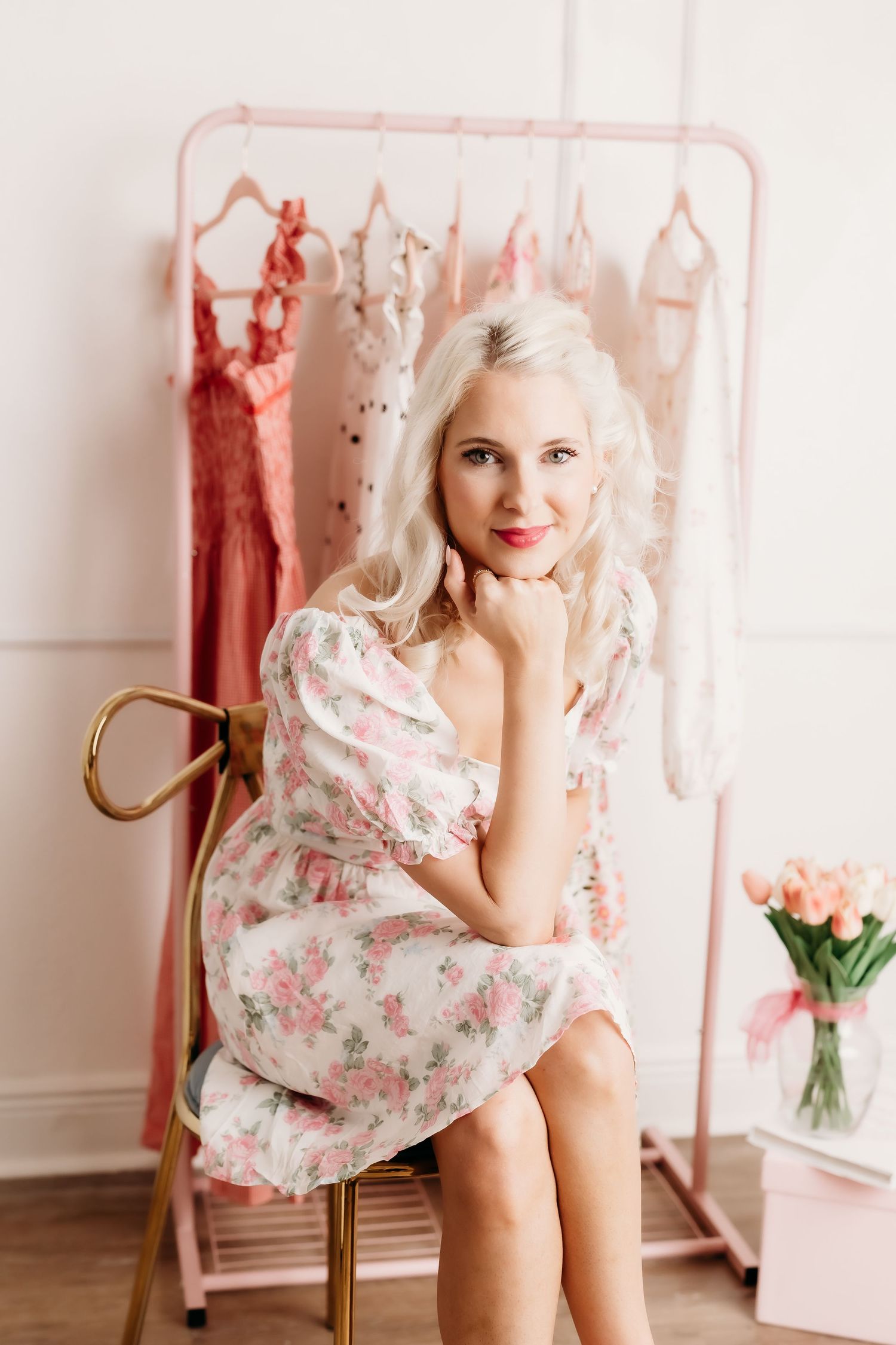 Blonde woman in floral dress sitting before pink clothing rack with tulips in bright white studio.
