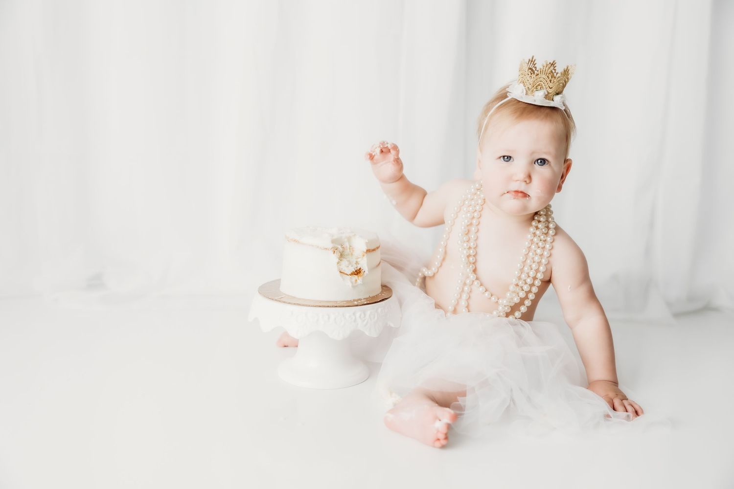 Baby girl in white tutu and gold crown sits beside a white first birthday cake on a white background.