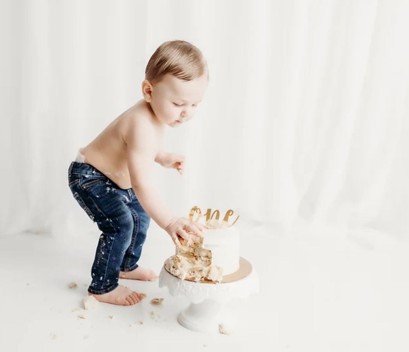 Toddler in jeans smashing a white first birthday cake with gold one topper.