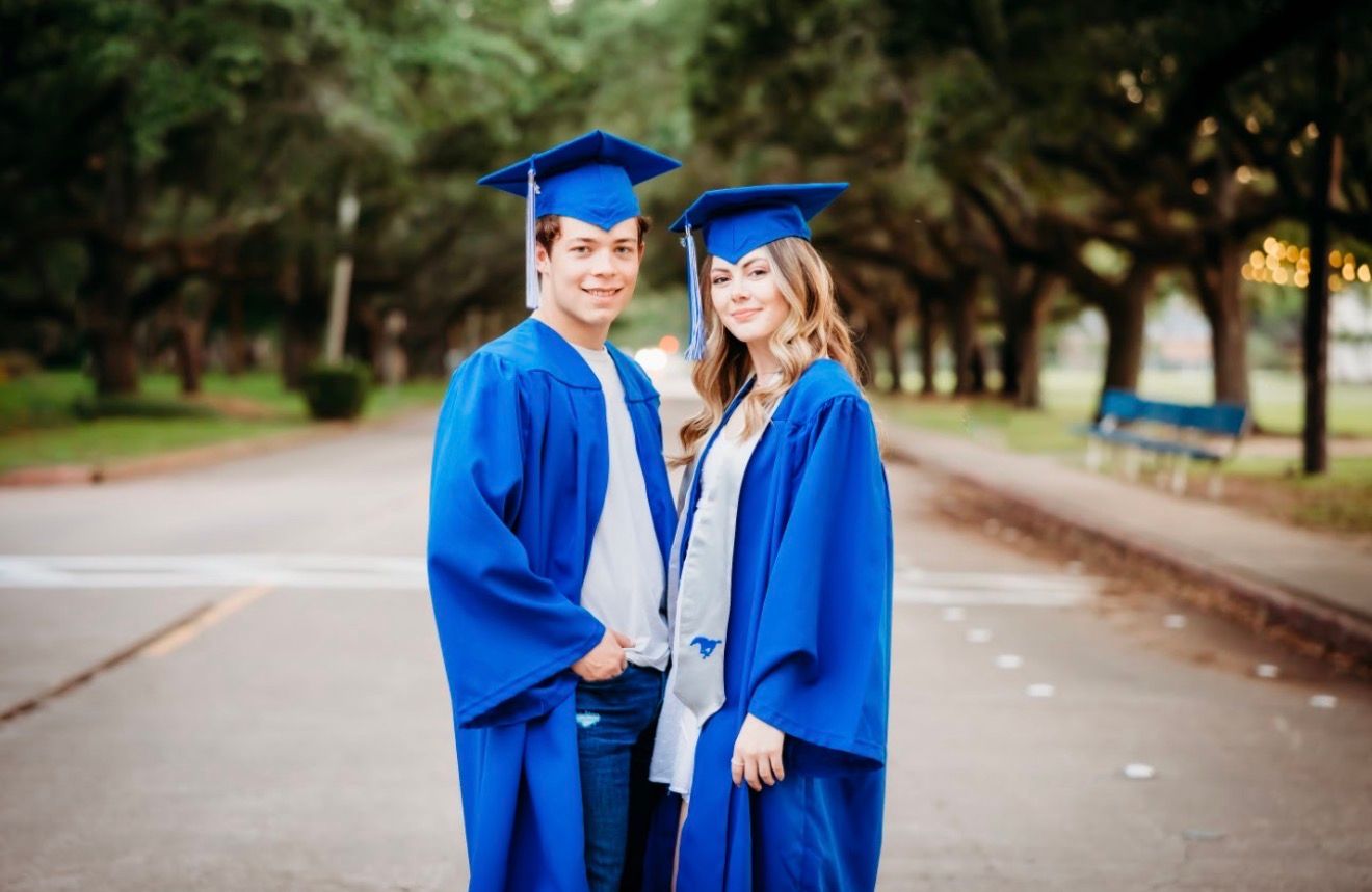 Two smiling graduates pose together in blue caps and gowns on a tree-lined path.