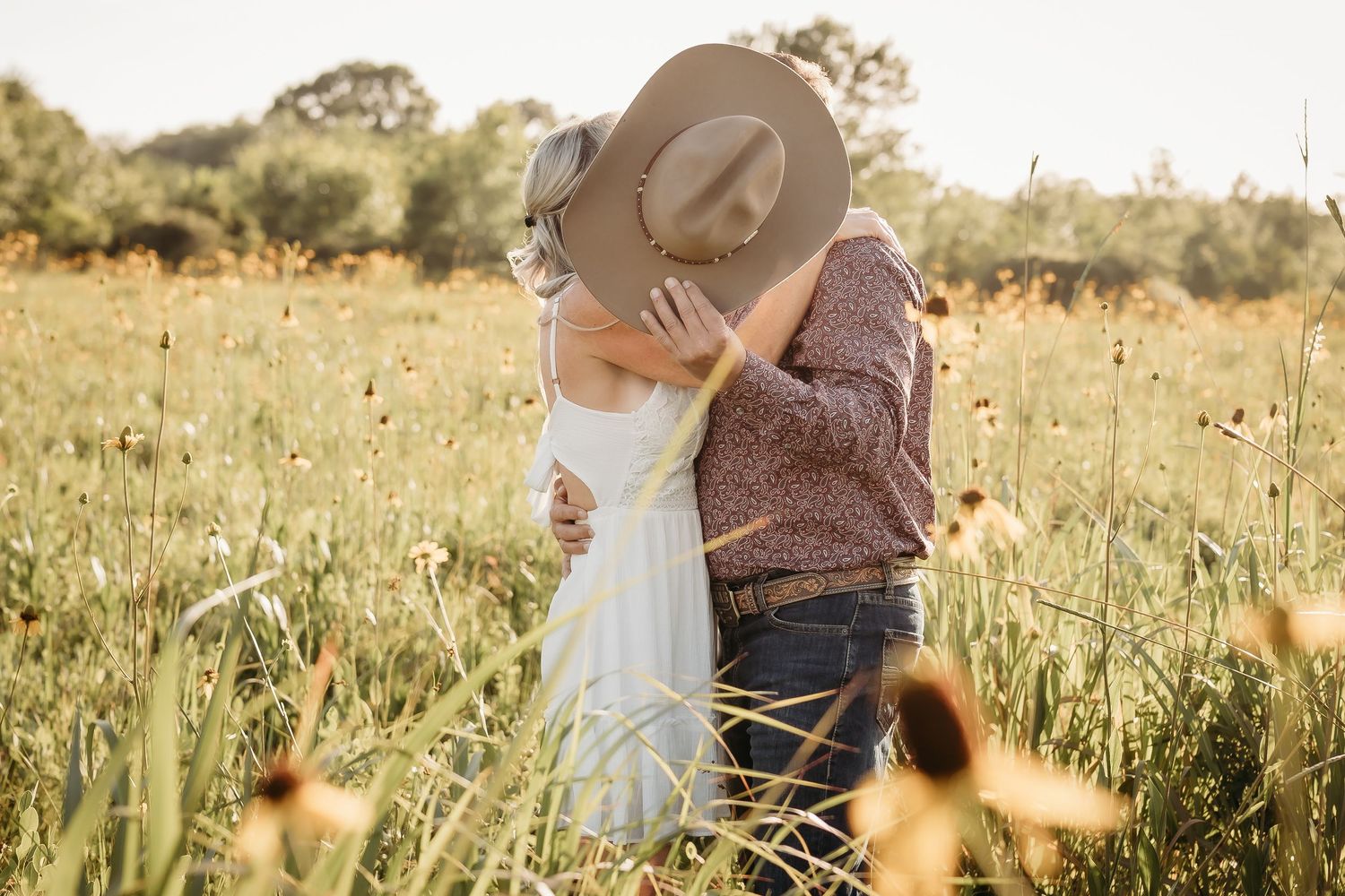 Couple sharing a romantic kiss hidden beneath a cowboy hat in a wildflower meadow.