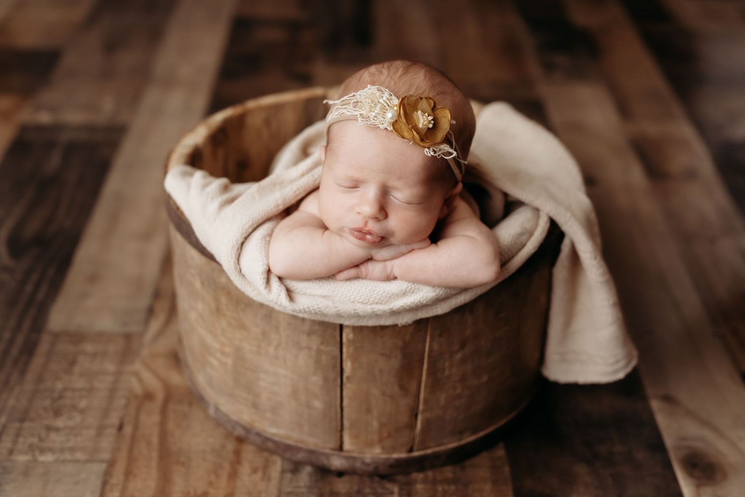 Sleeping newborn baby posed in rustic wooden bucket with cream wrap and gold floral headband.