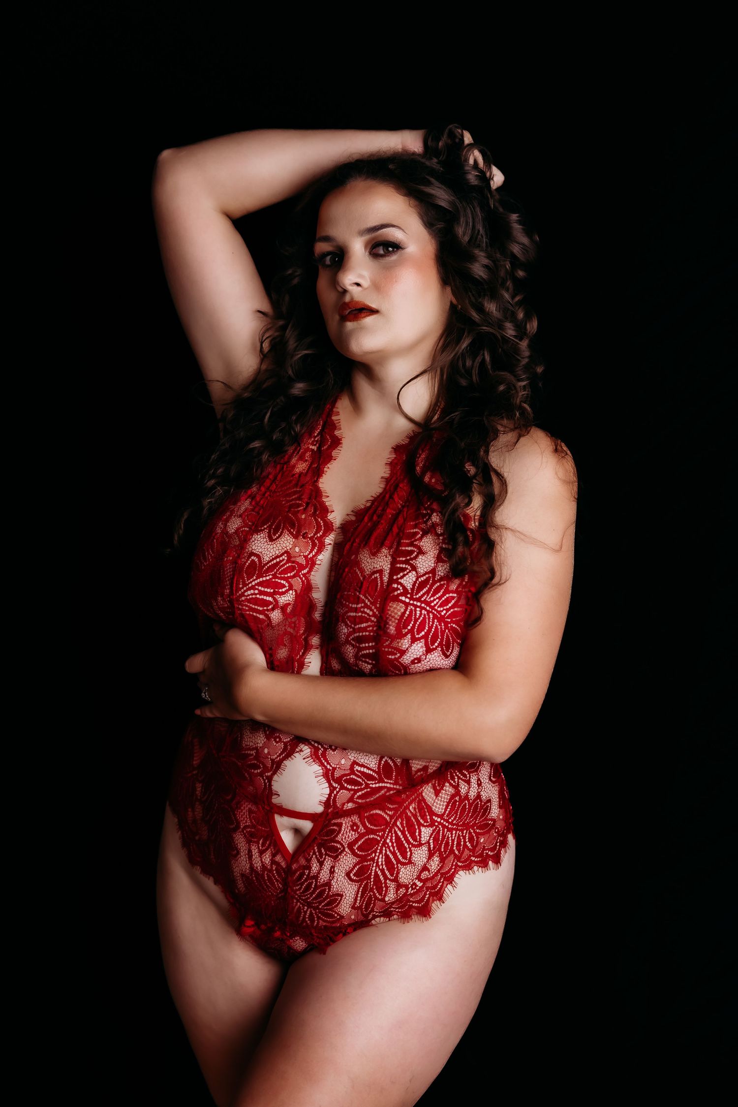 Curvy woman in red lace bodysuit posing confidently against a dark background in boudoir photo.