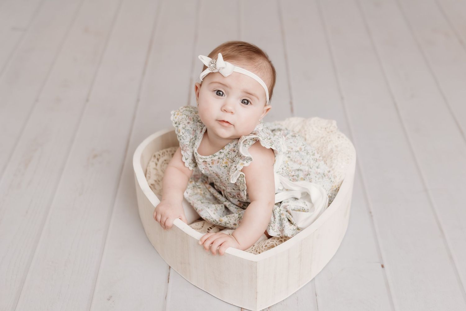 Adorable baby girl in lace romper sitting in wooden heart-shaped bowl on white floor.