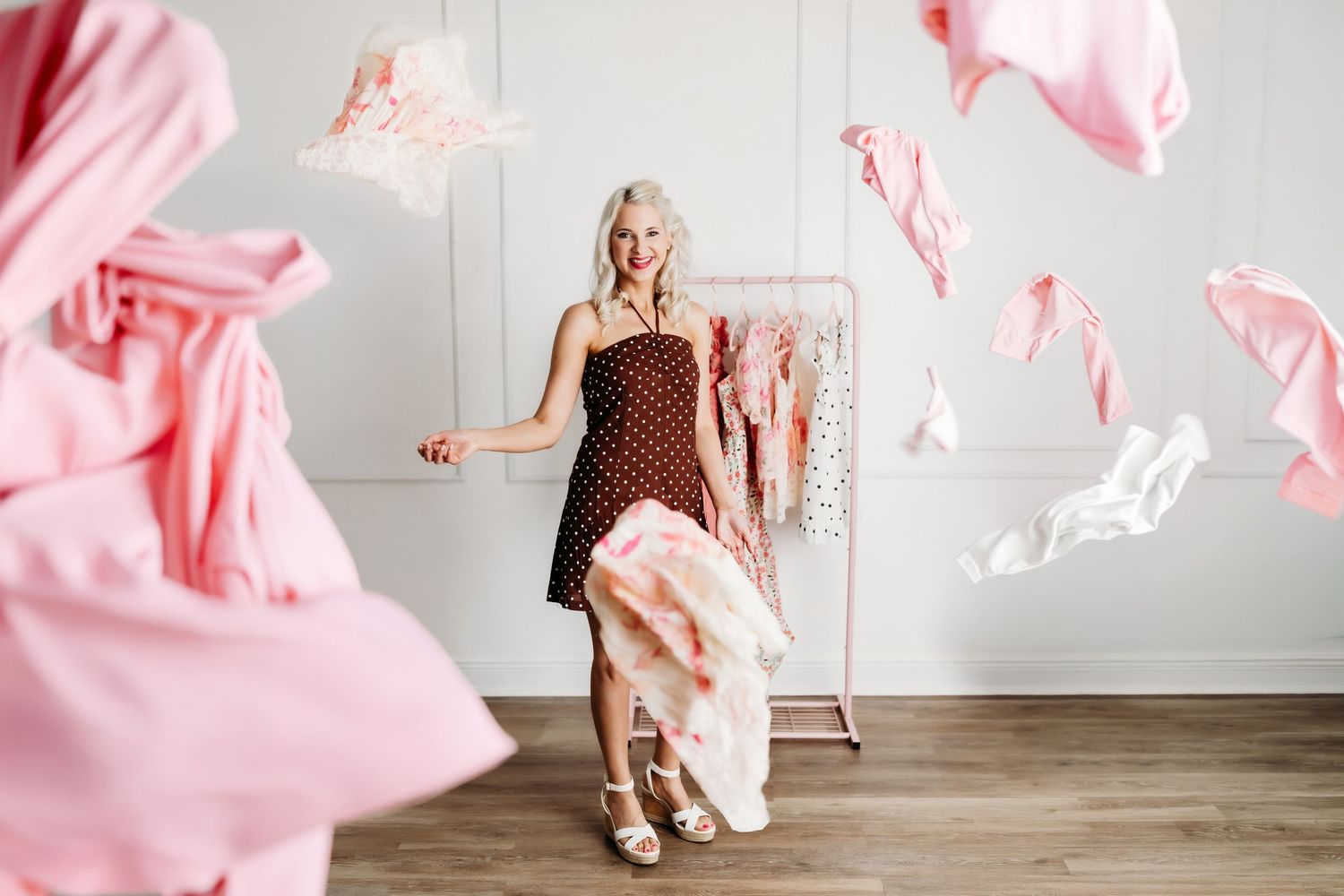 Blonde woman in polka dot dress surrounded by flying pink clothes near a clothing rack.