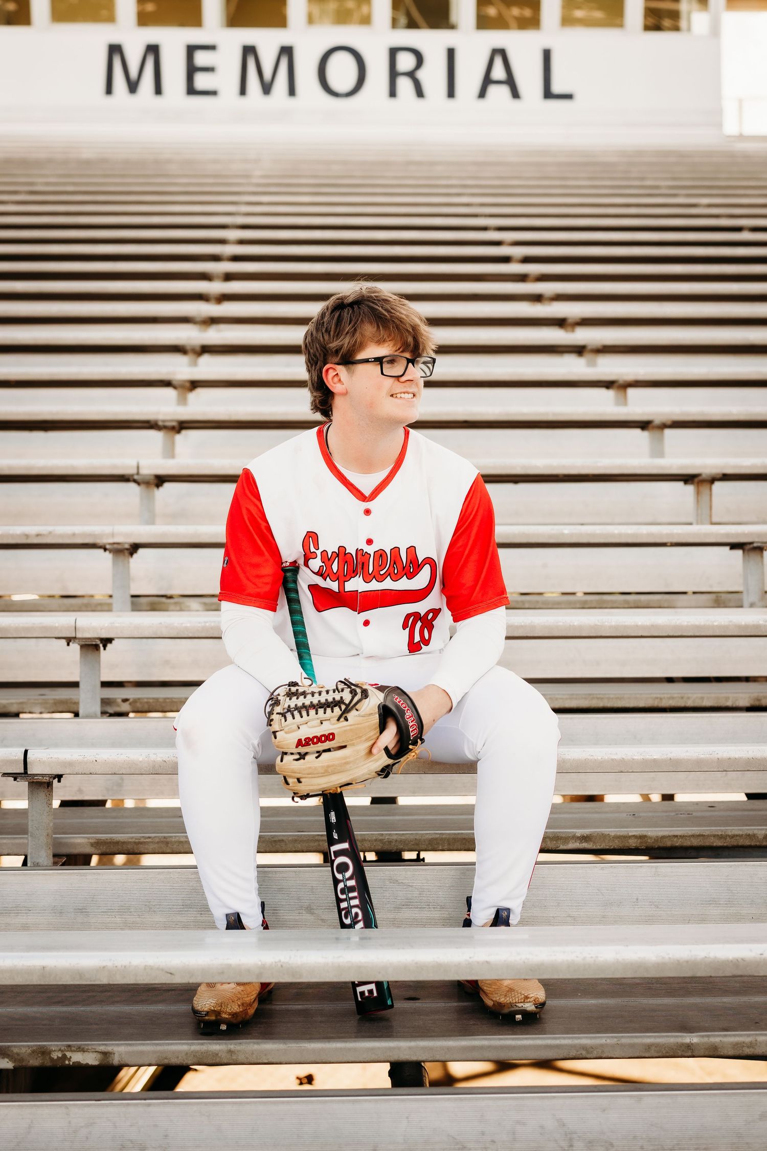 High school baseball senior in Express uniform sits in Memorial stadium bleachers with bat.