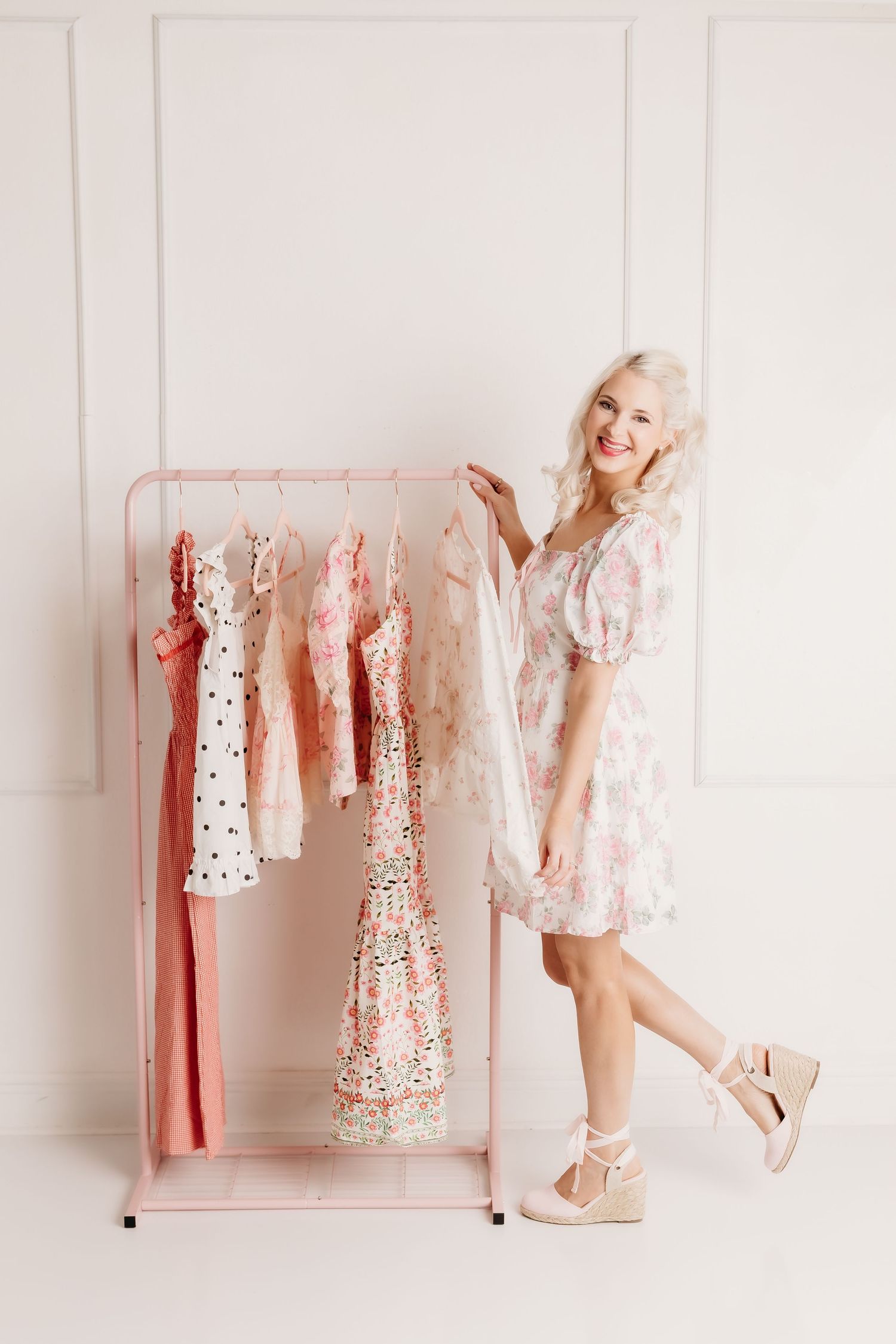 Smiling blonde woman in floral dress browsing pink clothing rack with pastel dresses.
