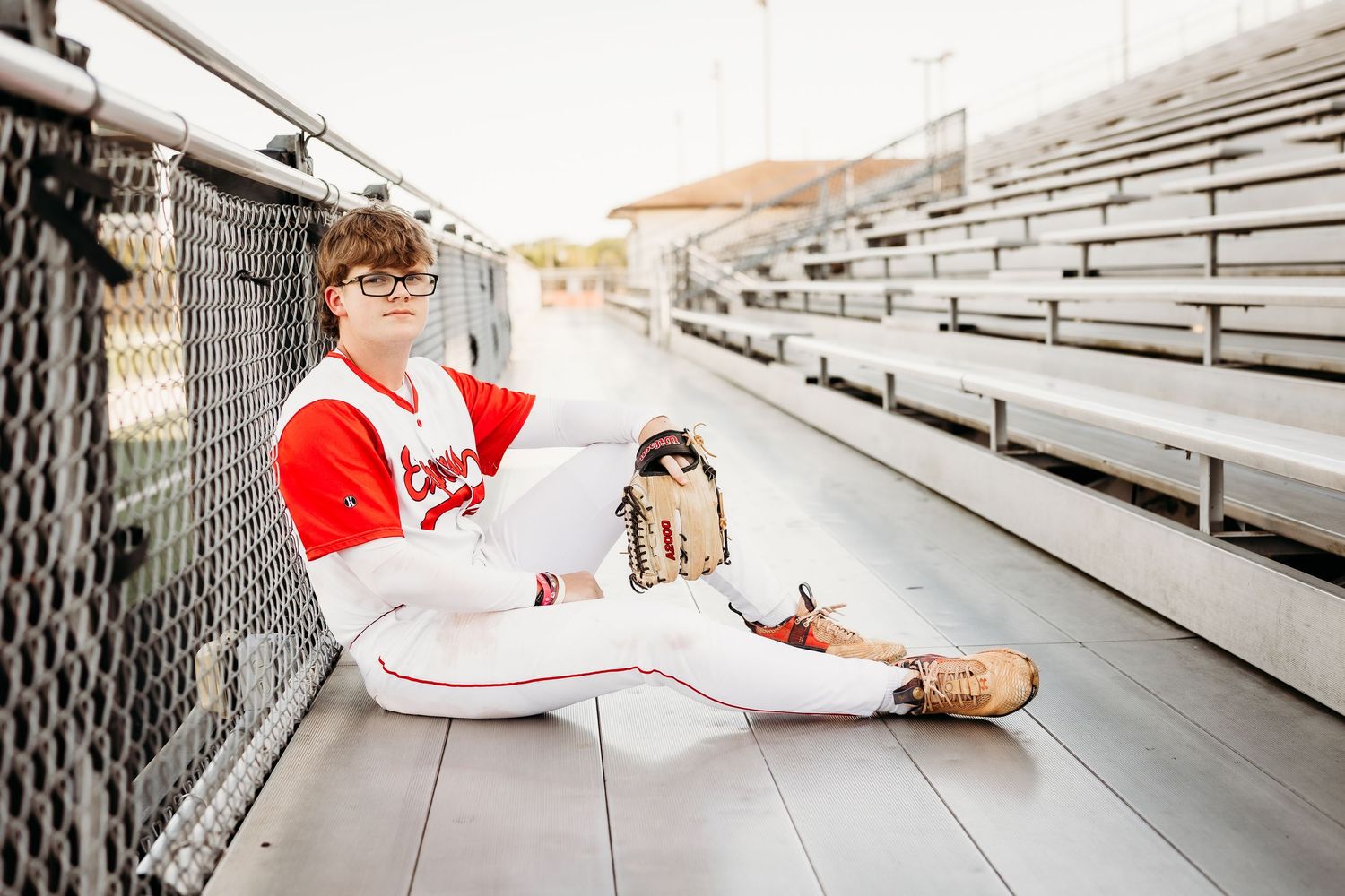 Baseball senior in Express uniform sits with glove and bat in stadium bleachers.