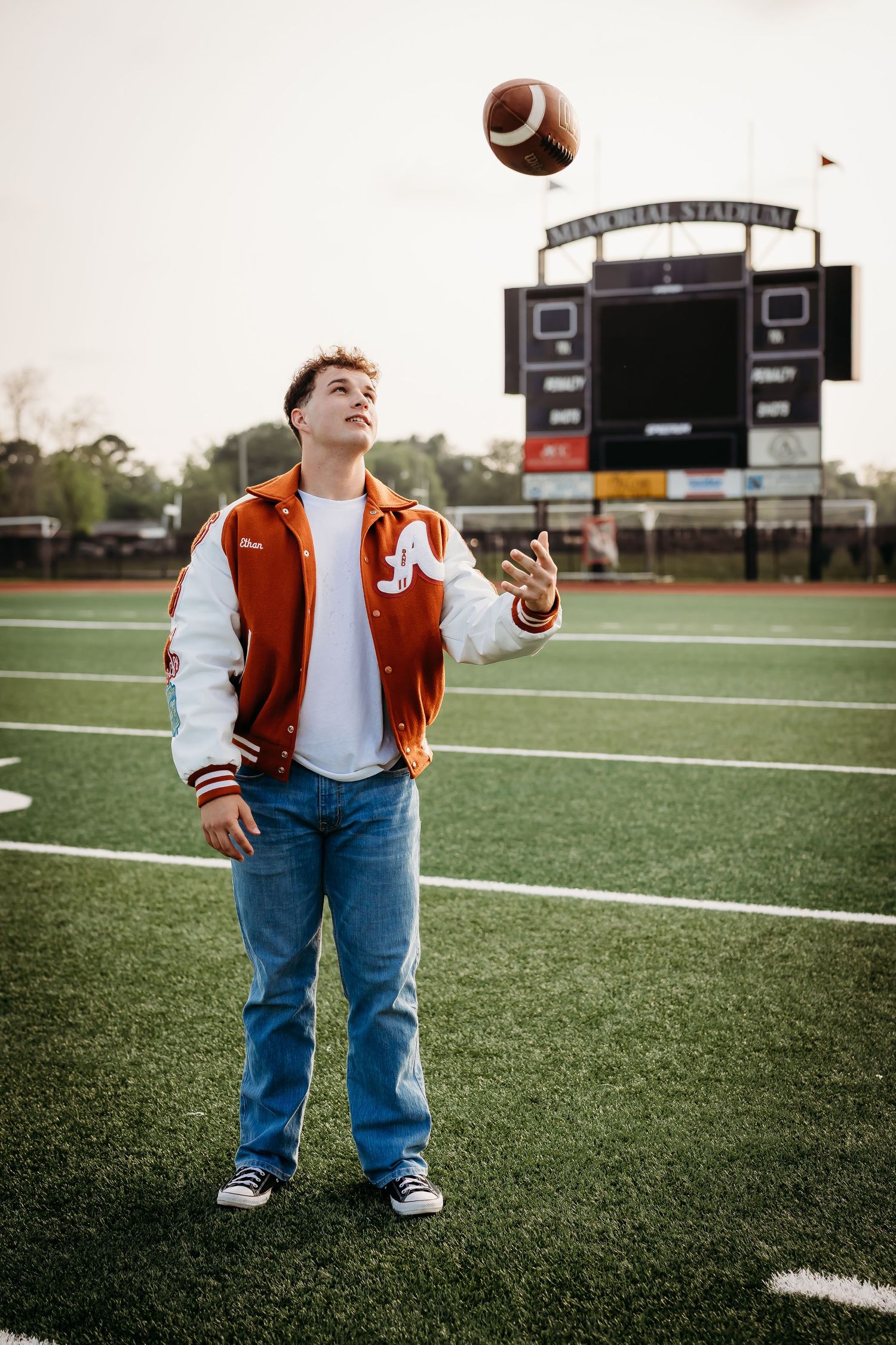 High school senior in orange letterman jacket tosses football on green turf field.